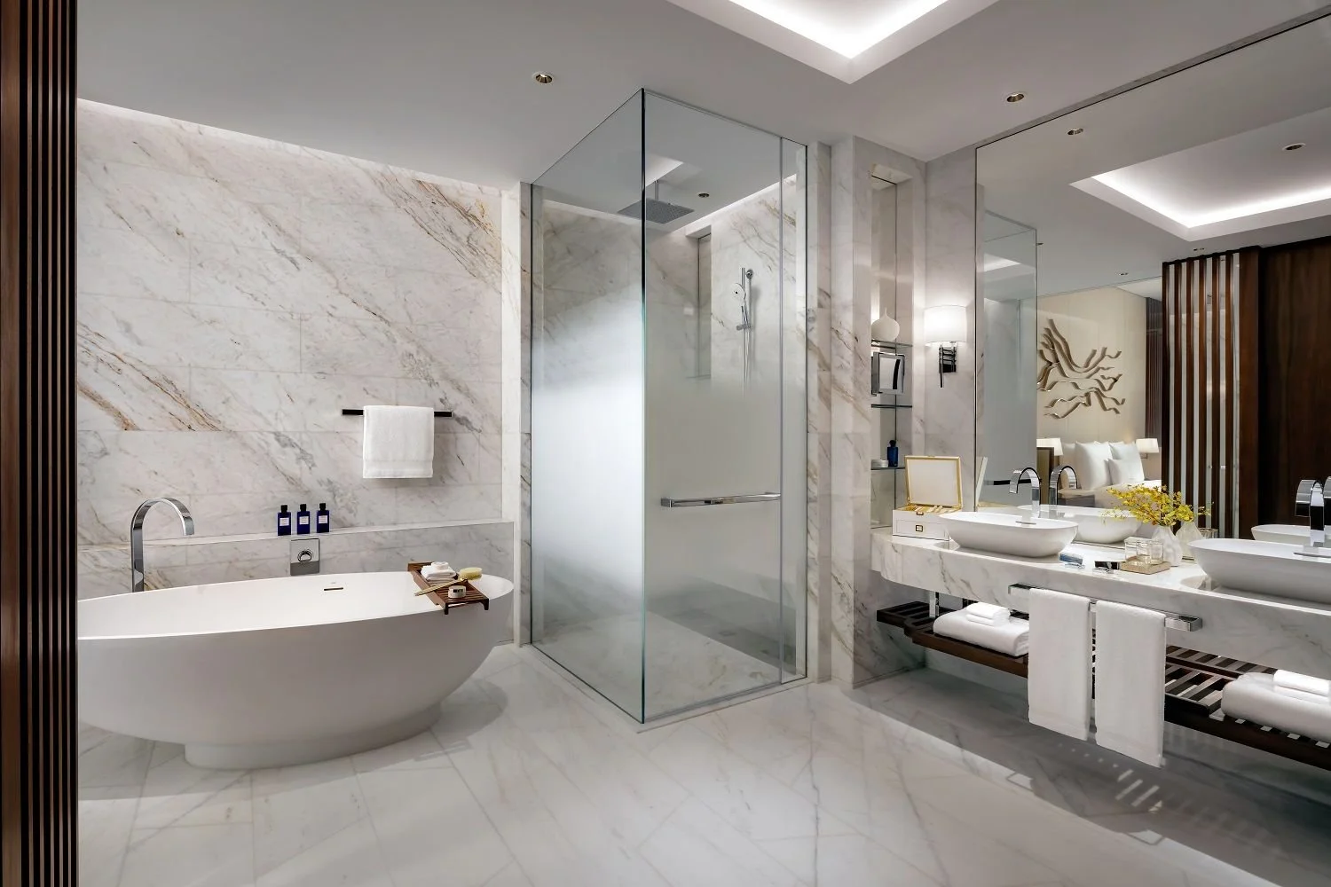 Luxurious bathroom with marble walls and floors, featuring a white freestanding bathtub with toiletries, a glass-enclosed shower, a double sink vanity with towels and decorative items, and a view into a bedroom with bed and wall art.