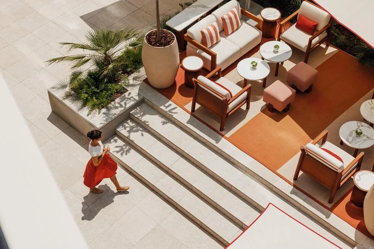 An aerial view of a woman in a white top and red skirt walking down steps towards a creatively arranged outdoor patio with white seating, armchairs, small tables, and potted plants.