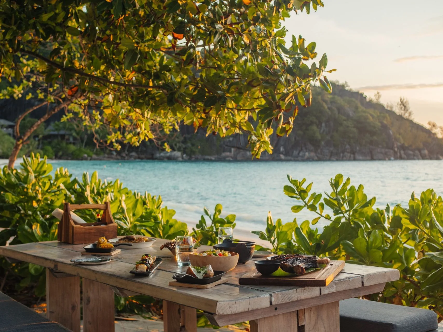 Dinner table set with plates of food on a wooden table outdoors by a beach, with greenery and a hillside in the background at sunset.