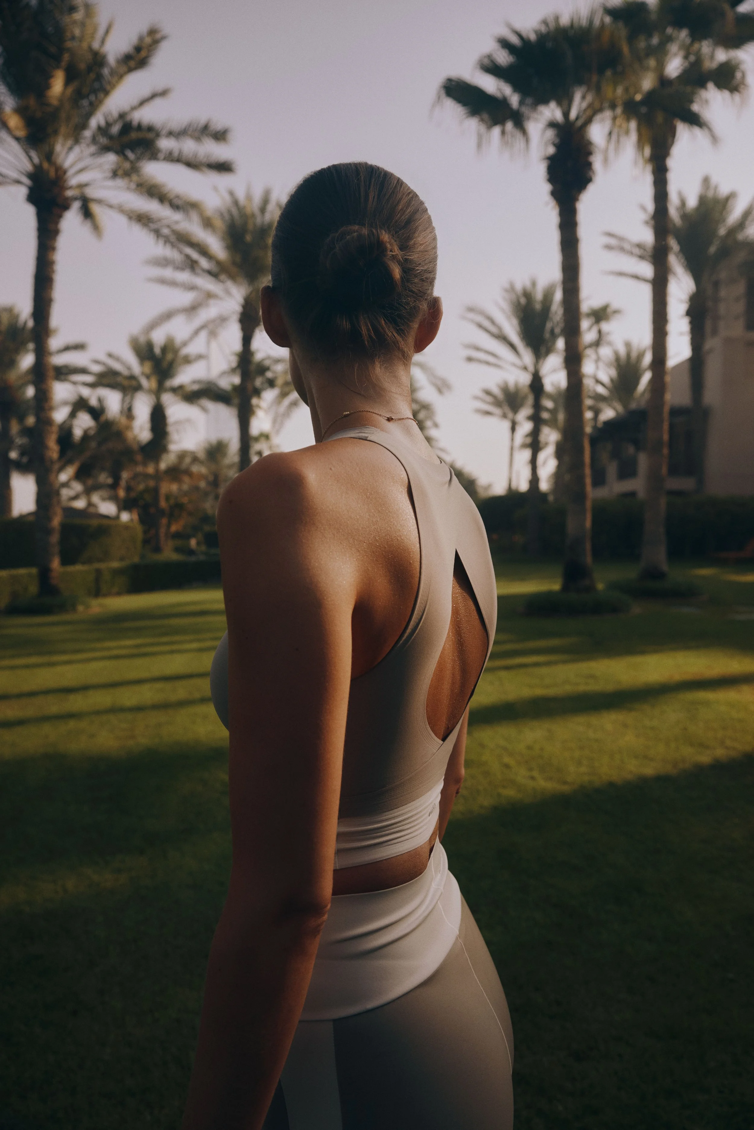 A woman with her hair in a bun wearing athletic clothing outdoors among palm trees during sunset.