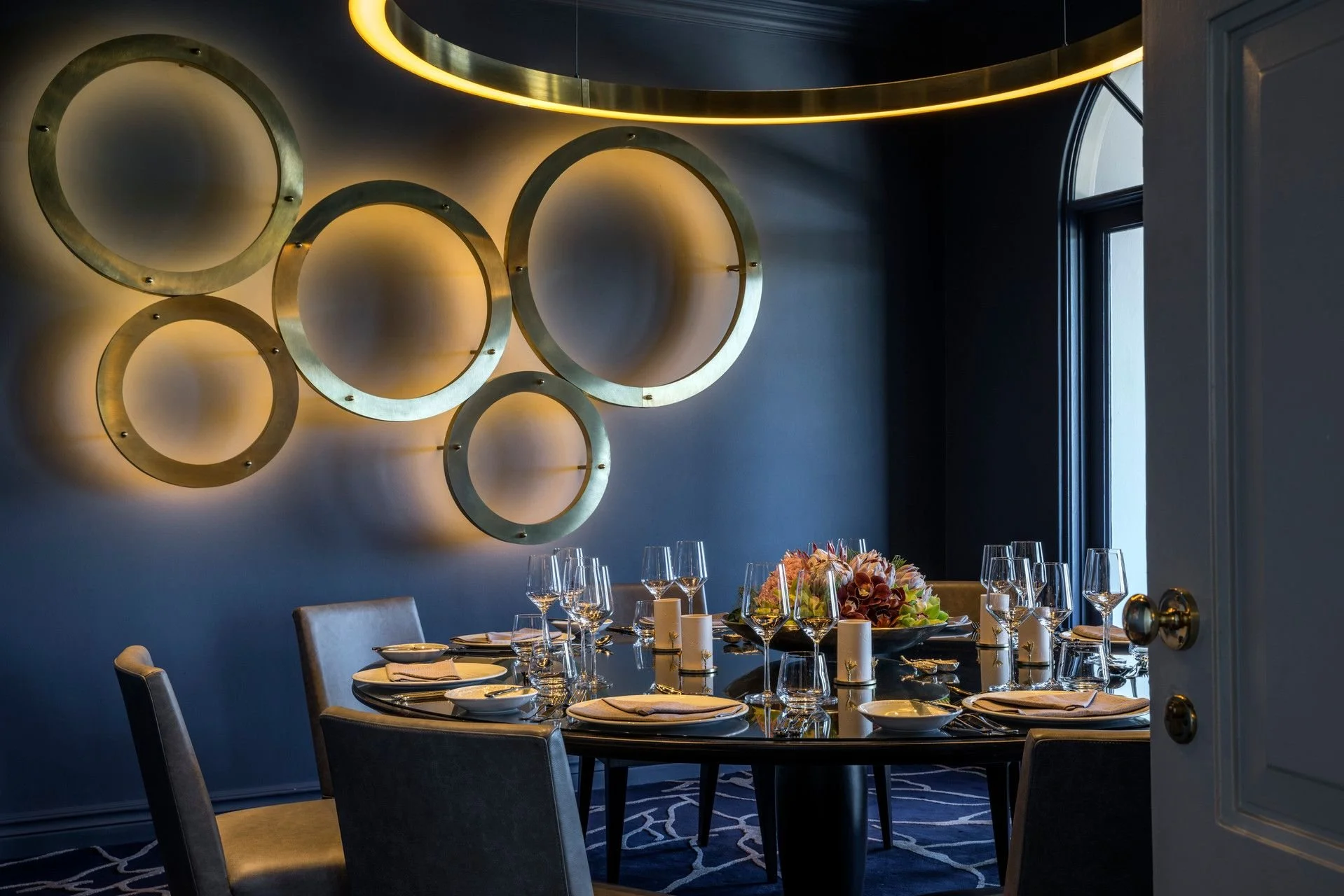 A dining room with a round table set for a formal meal, decorated with a floral centerpiece, glasses, plates, and candles. The wall behind features modern circular gold wall art with backlighting, and there is a window with dark frames on the right side.