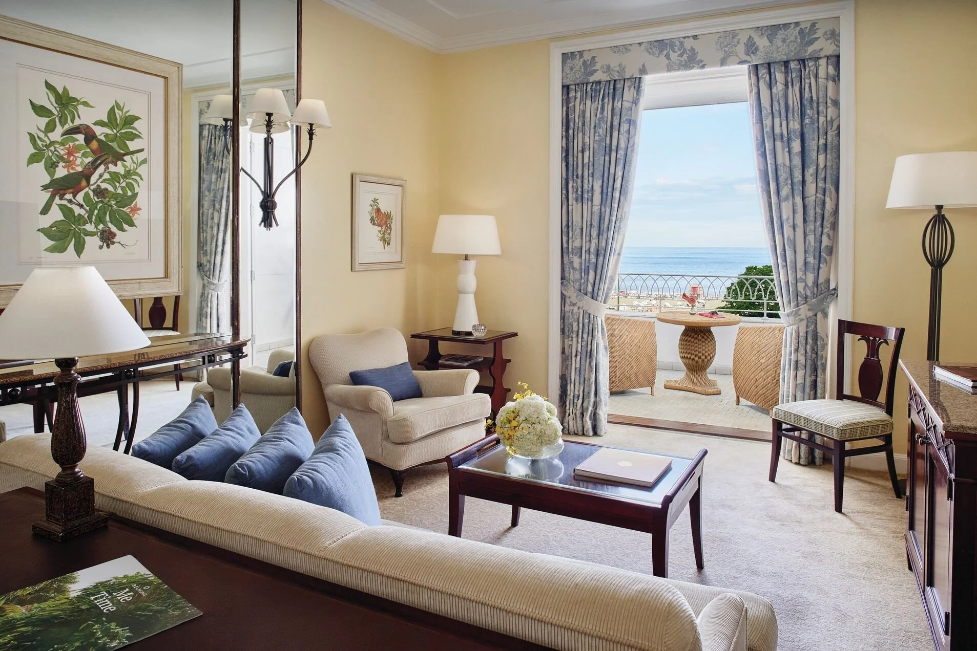 Living room with yellow walls, blue curtains, and a balcony view of the ocean, featuring a cream armchair, a glass coffee table with a flower arrangement, and blue cushions on a beige sofa.