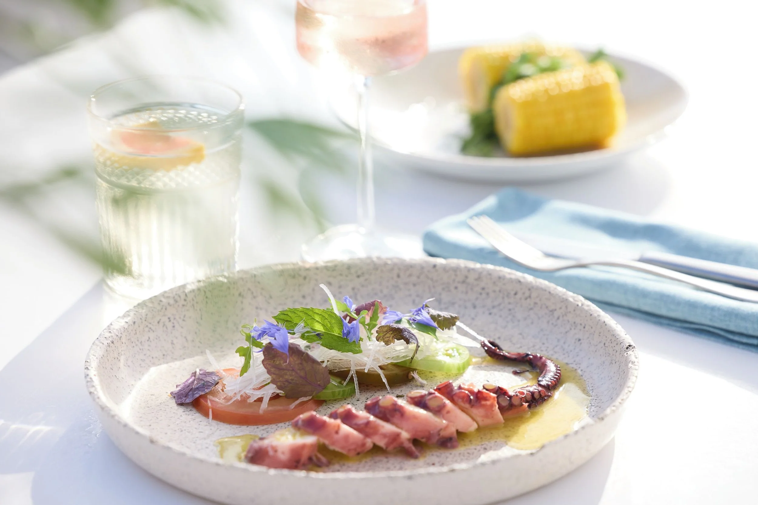 A plate of thinly sliced smoked salmon garnished with fresh herbs and edible flowers, with a few slices of cucumber and microgreens on top. In the background, there are glasses of sparkling water with lemon slices, a glass of rosé wine, and a plate of grilled corn with herbs.