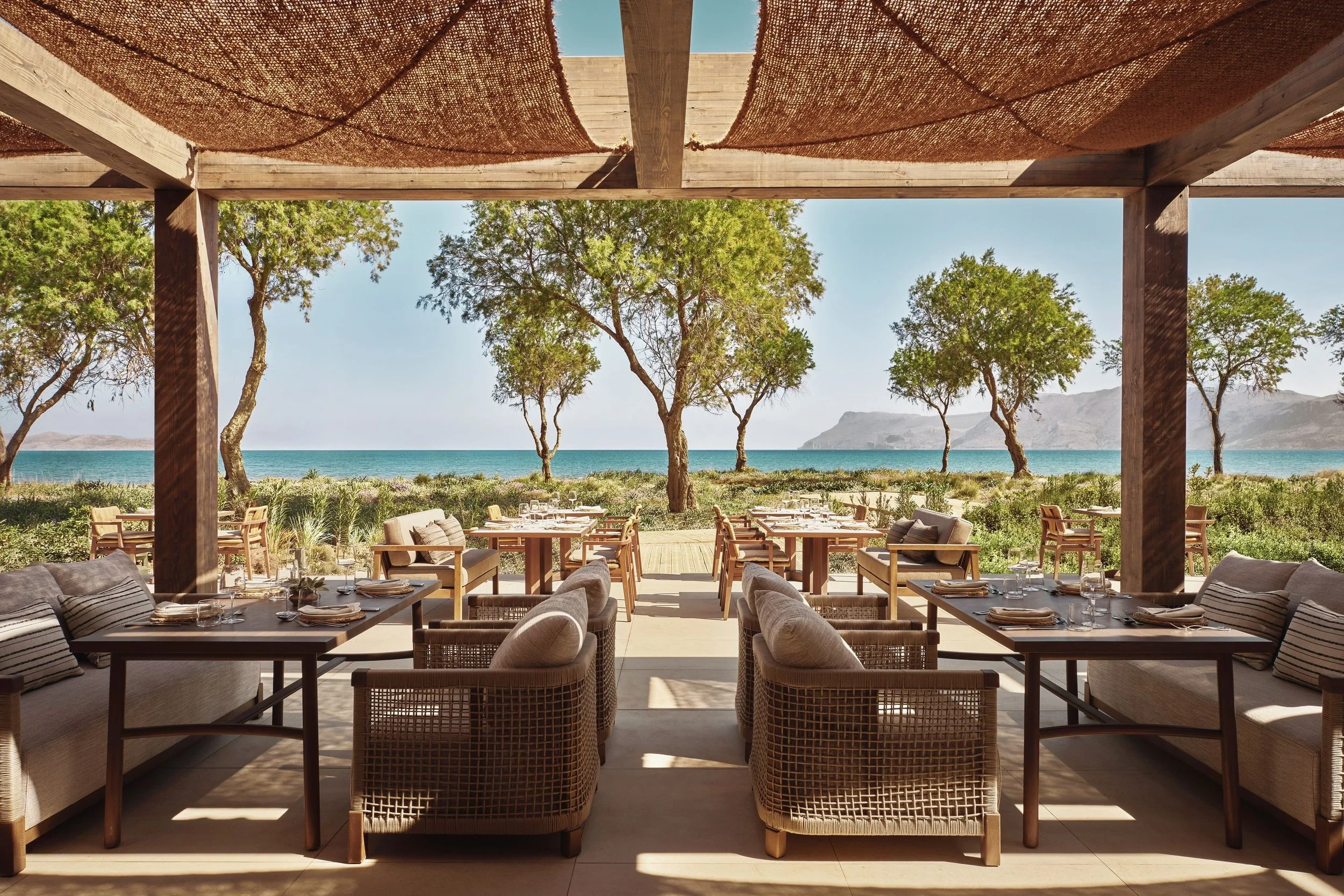 Outdoor dining area with tables and chairs, overlooking a beach and ocean with trees and mountains in the background.
