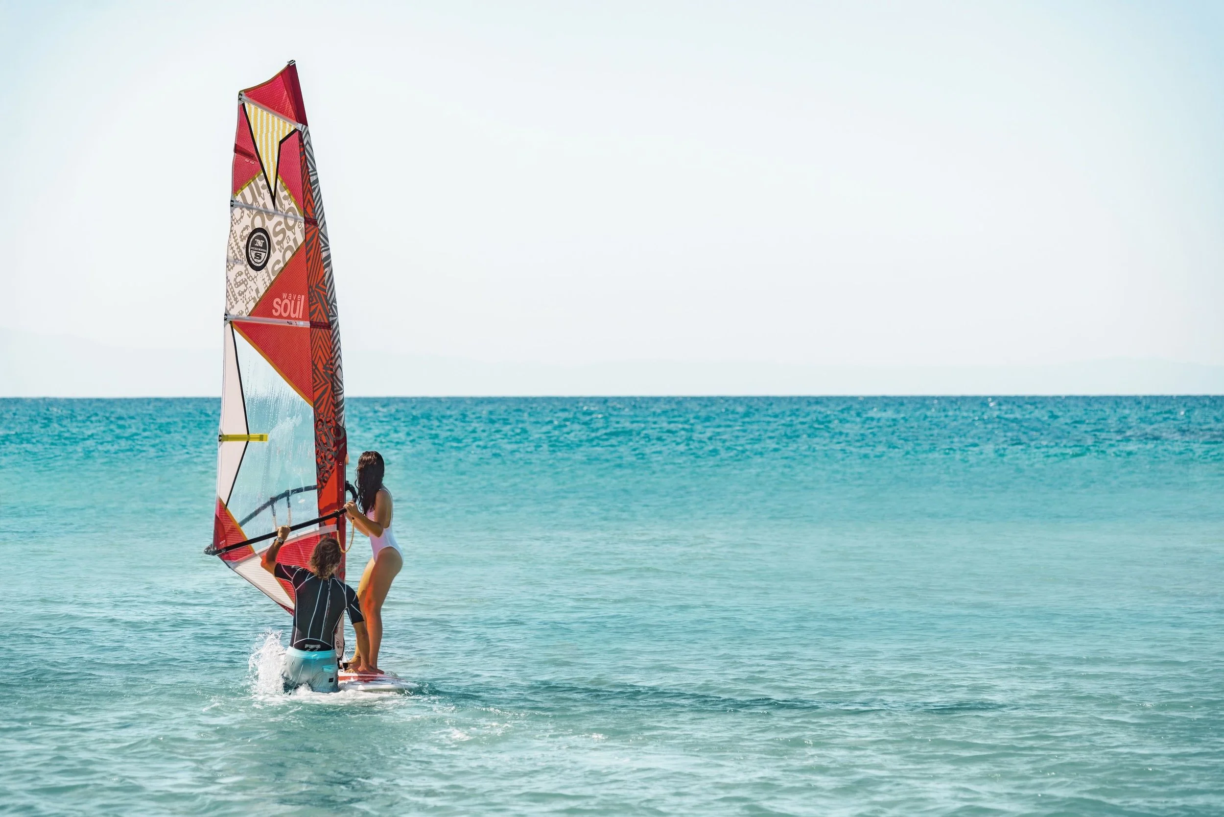 Two people windsurfing on the ocean with a colorful sail, near the shore with turquoise water and a clear sky in the background.