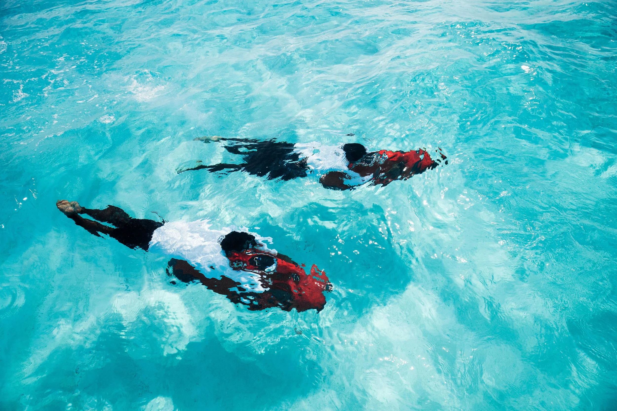 Two individuals swimming in a clear blue swimming pool, wearing life jackets.