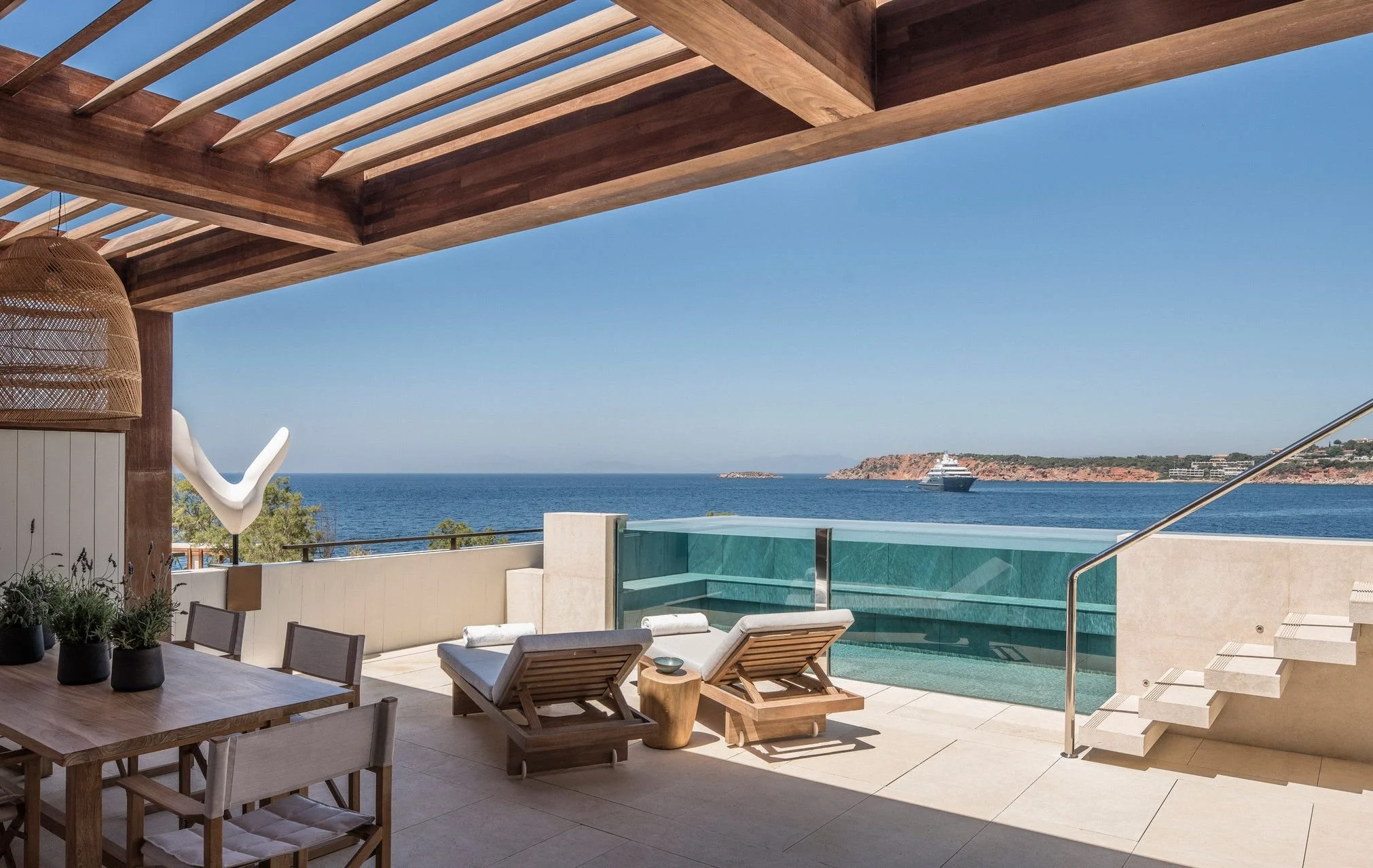 A modern outdoor patio with a view of the ocean, featuring a wooden pergola, lounge chairs, a swimming pool, and a cruise ship in the water.
