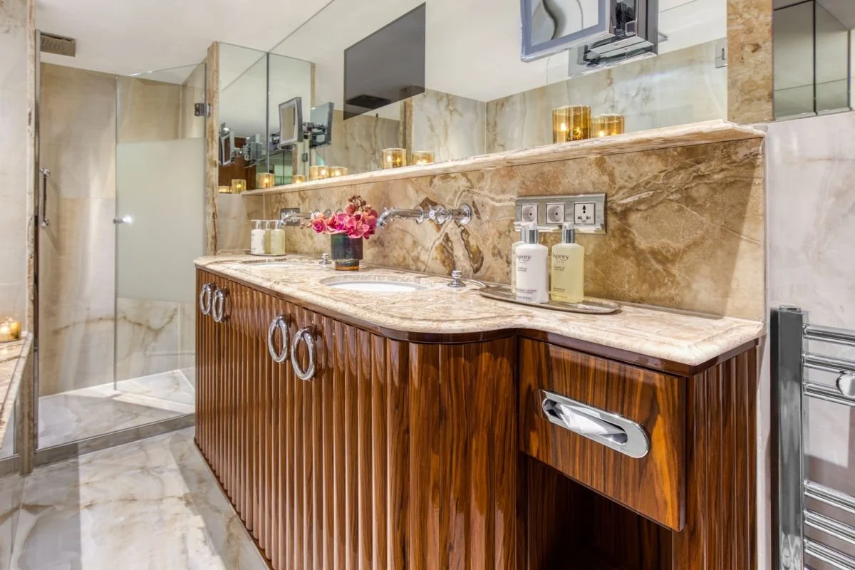 A modern bathroom with a marble countertop, wooden cabinet, flowers, soap dispensers, and golden candles, with a glass shower enclosure and large mirror.