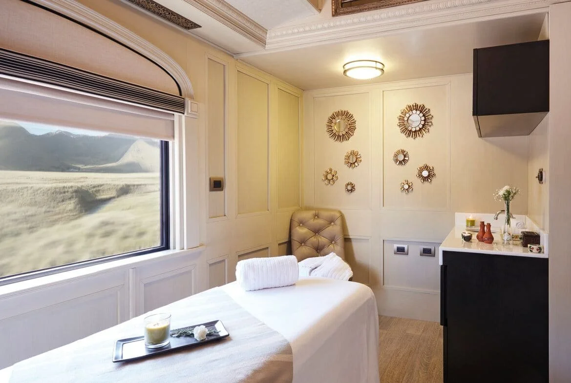 A cozy, well-decorated room with a massage or treatment bed, a window with a view of mountains and land, and a wall adorned with decorative mirrors. The room has warm lighting and a small counter with a vase of flowers, candles, and other decor items.