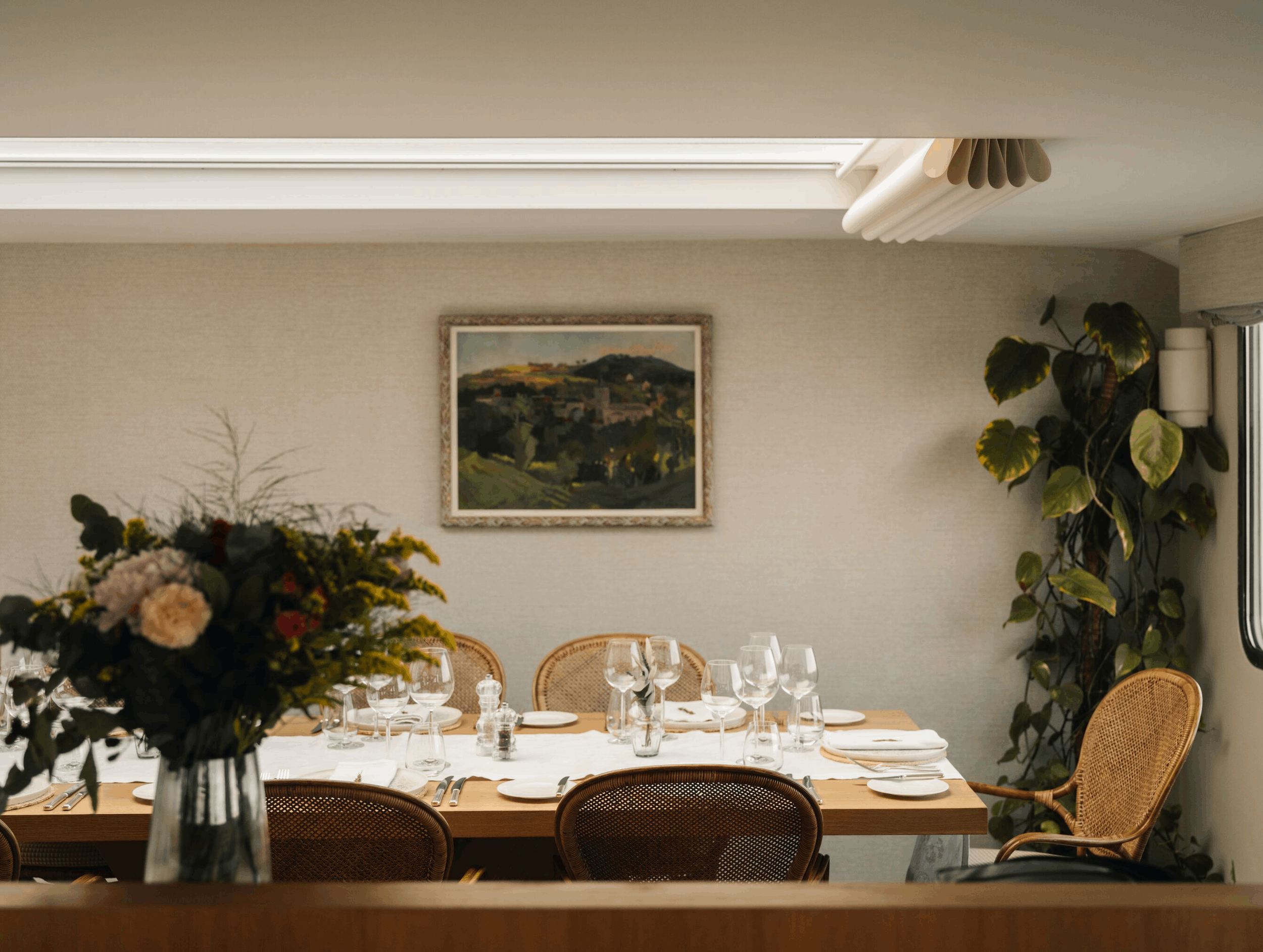 A dining table set for a formal meal with wine glasses, plates, and cutlery, decorated with a large bouquet of flowers, in a room with framed landscape painting, green plants, and a beige wall.