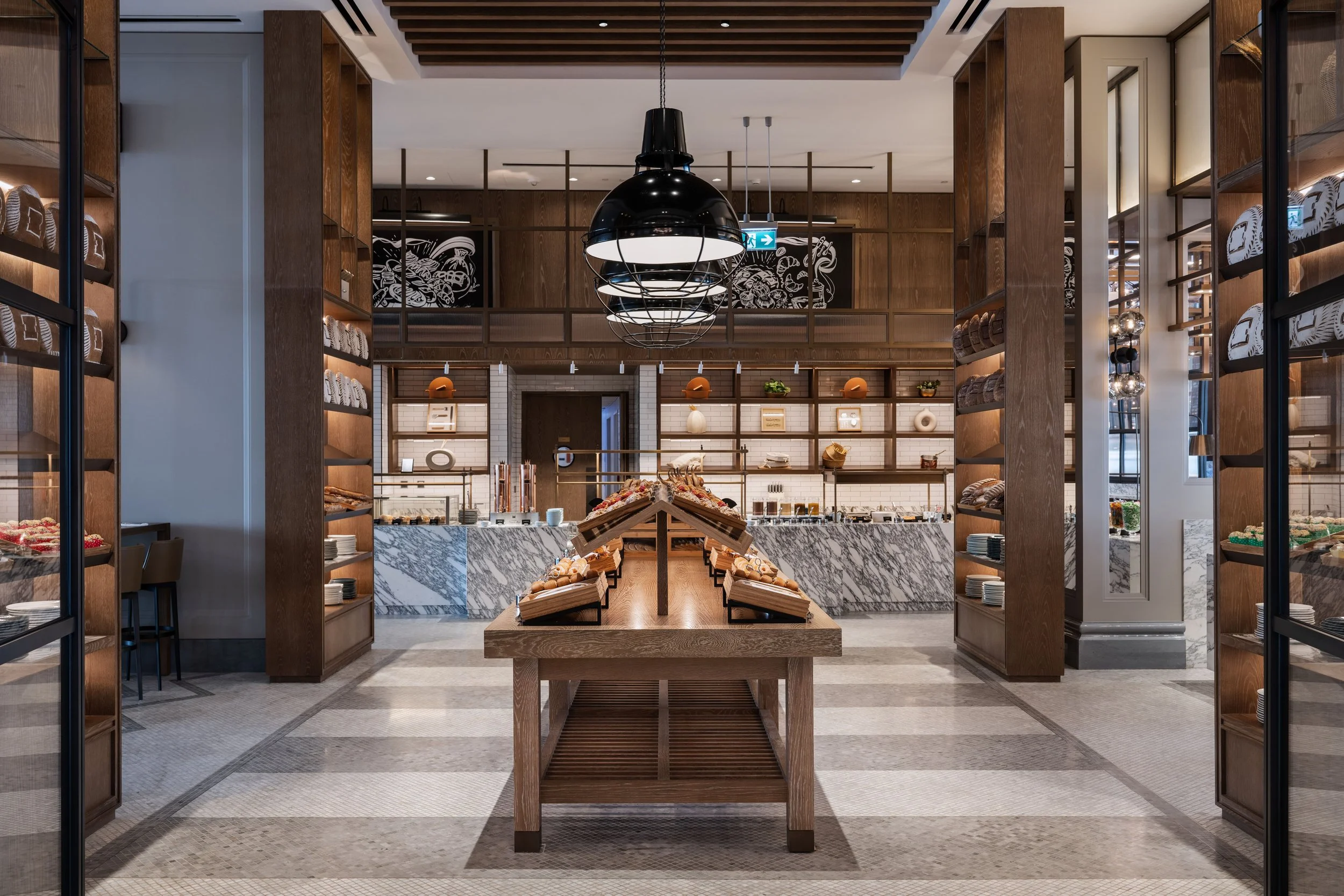 Interior view of a modern bakery or cafe with wooden shelving, light fixtures, and a marble-topped counter displaying baked goods.