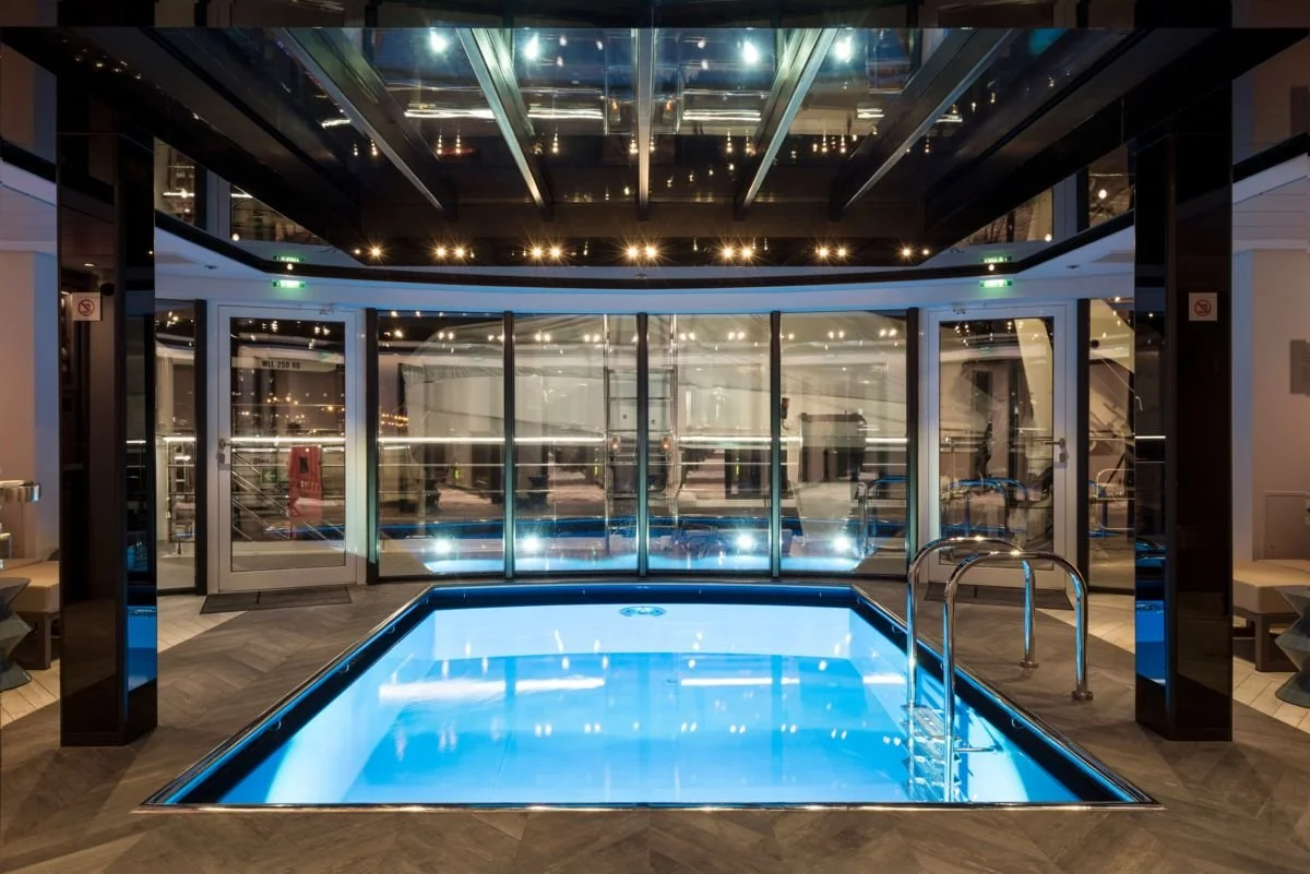 Indoor hotel or spa swimming pool with illuminated blue water, glass walls, and stainless steel handrails.