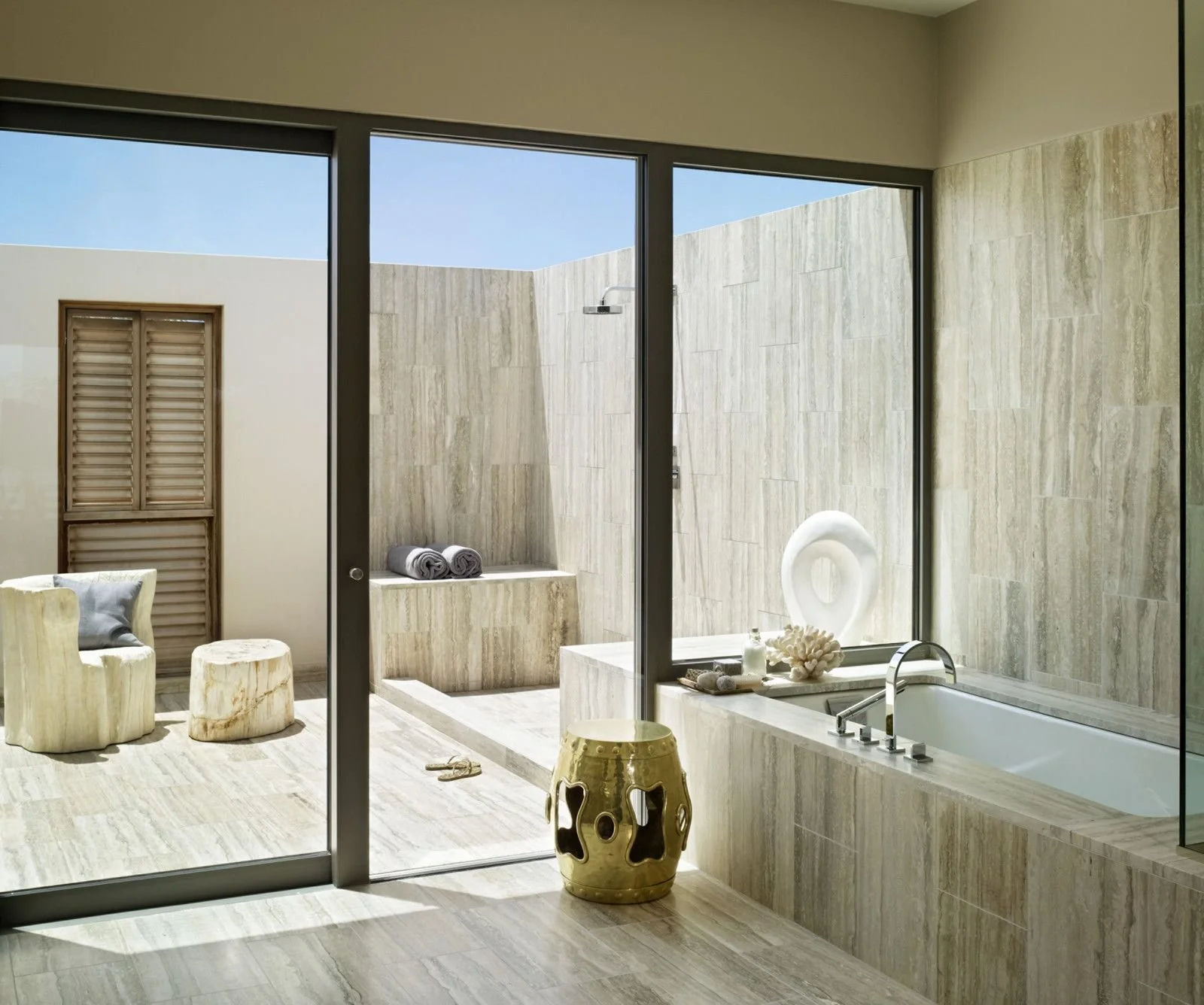 Modern bathroom with a large glass window, a bathtub, stone decor, and outdoor seating with wooden stools and rolled towels.