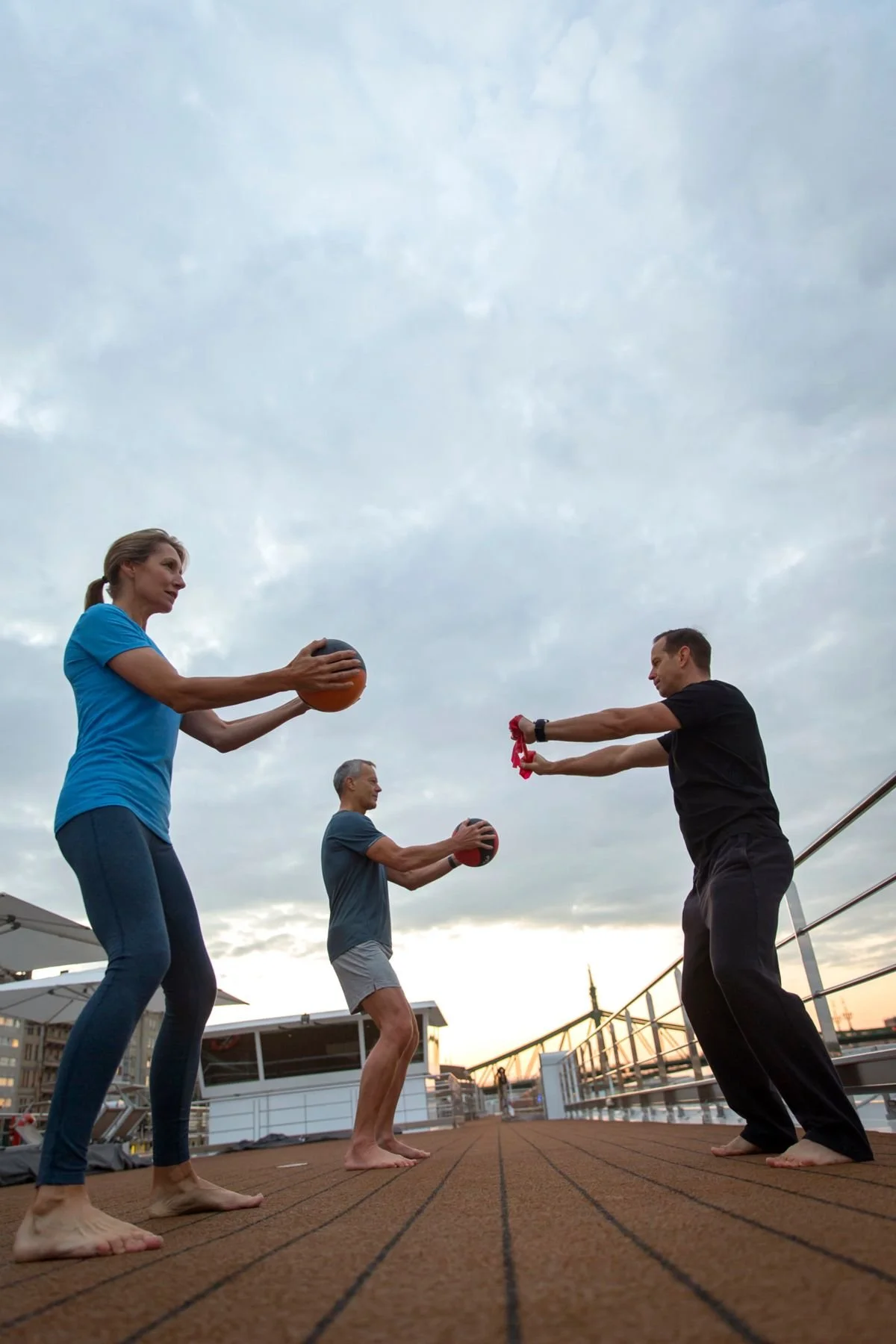 Three people doing outdoor fitness training with medicine balls on a boat deck during sunset.