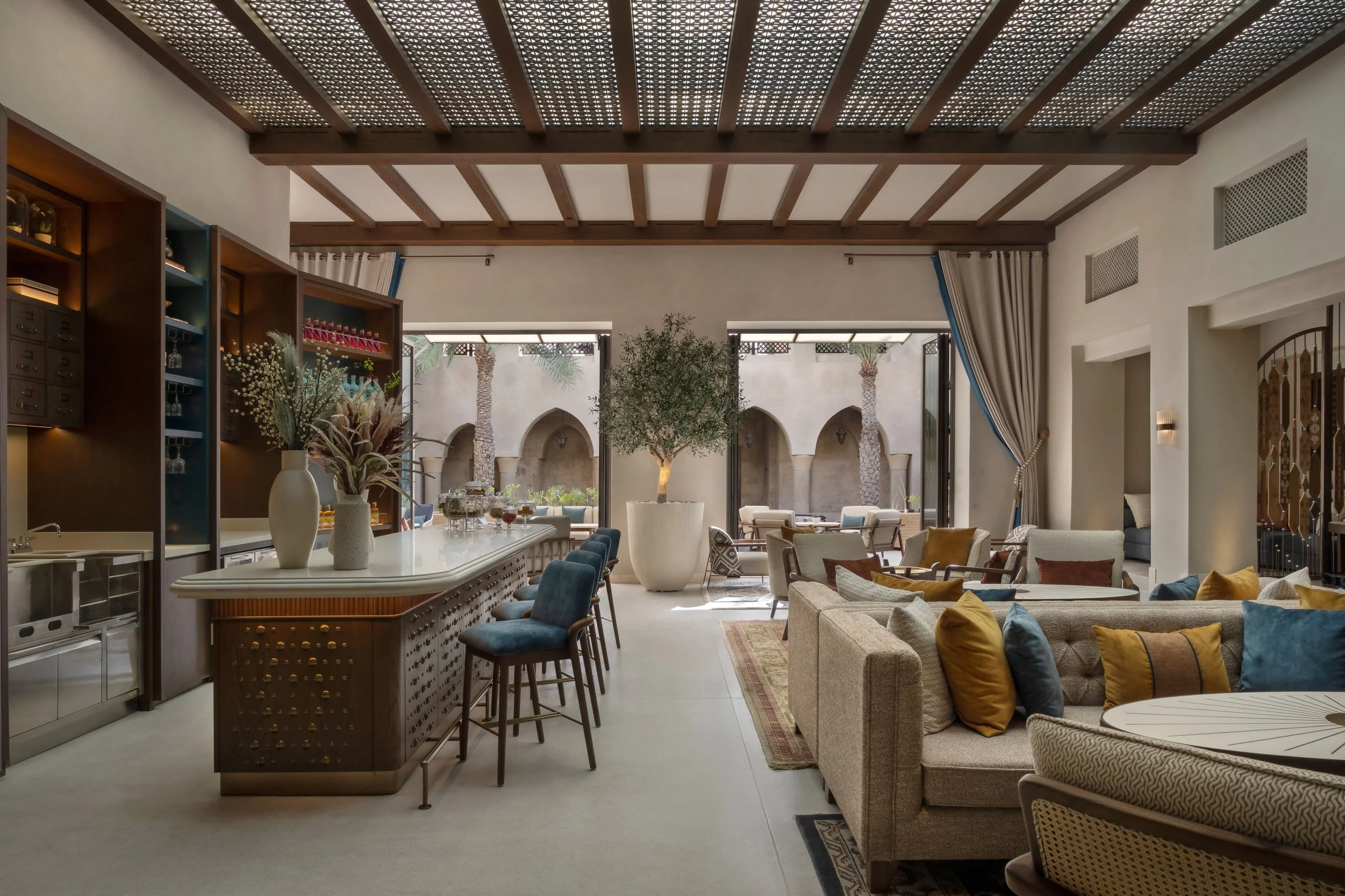 Interior of a spacious, modern living room with a high wooden ceiling, large windows, and multiple seating areas with sofas and chairs, decorated with cushions and plants.