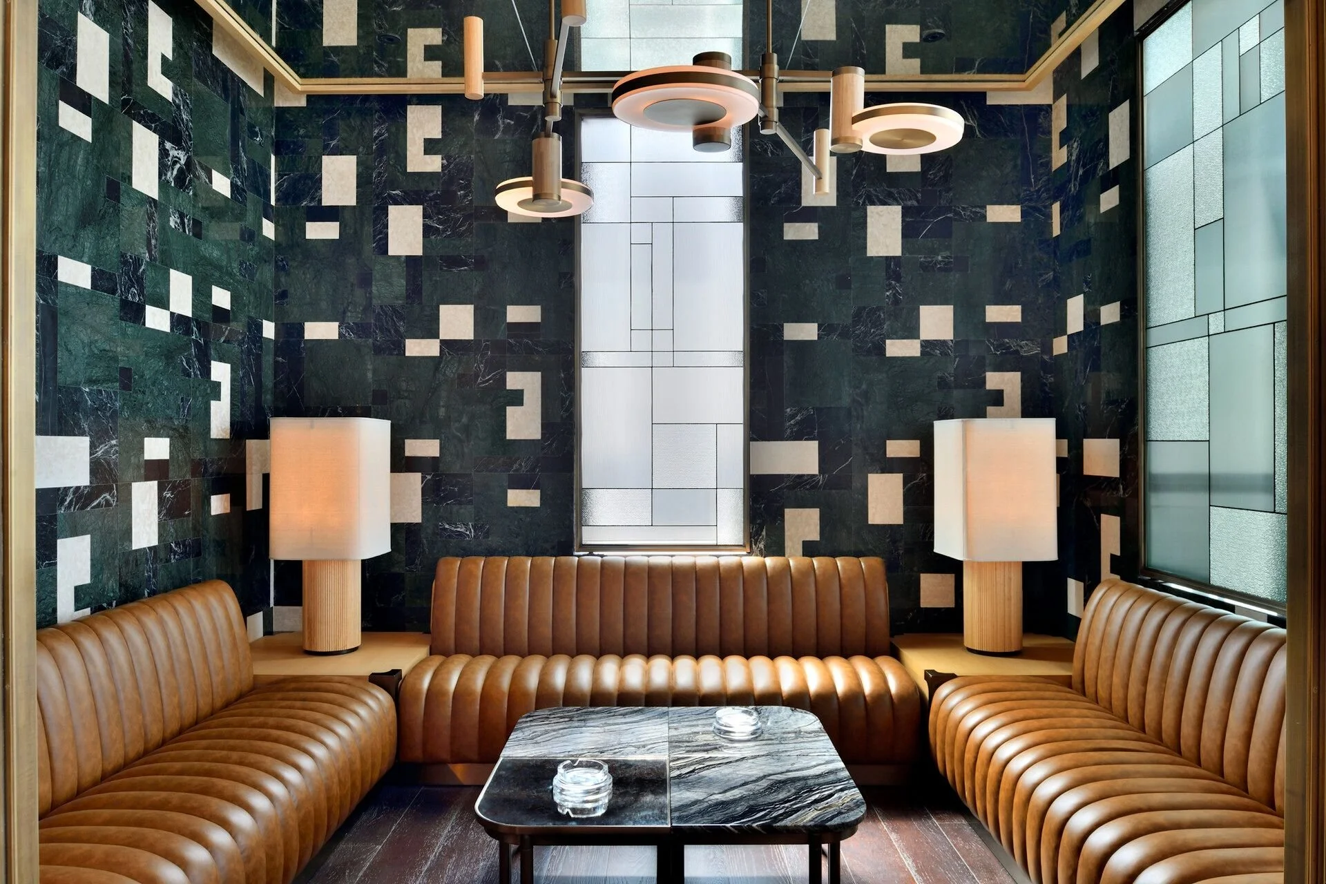 A modern lounge area featuring two tufted leather sofas facing each other with a marble-top coffee table in between. The walls are decorated with dark green and black marble tiles with white accents. There are two white table lamps with fabric shades on either side of the sofas. Suspended from the ceiling are three contemporary pendant lights with round and cylindrical shapes.