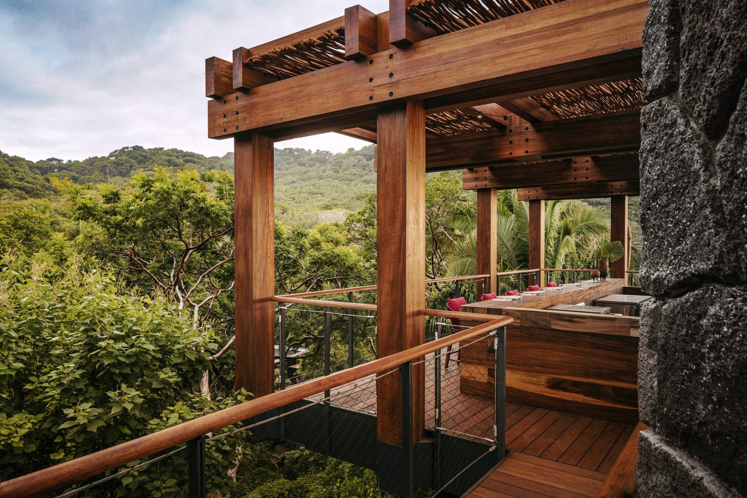 A wooden balcony overlooking lush green trees and hills in the background, with a dining area set with plates and glasses, and a rugged stone wall on the side.