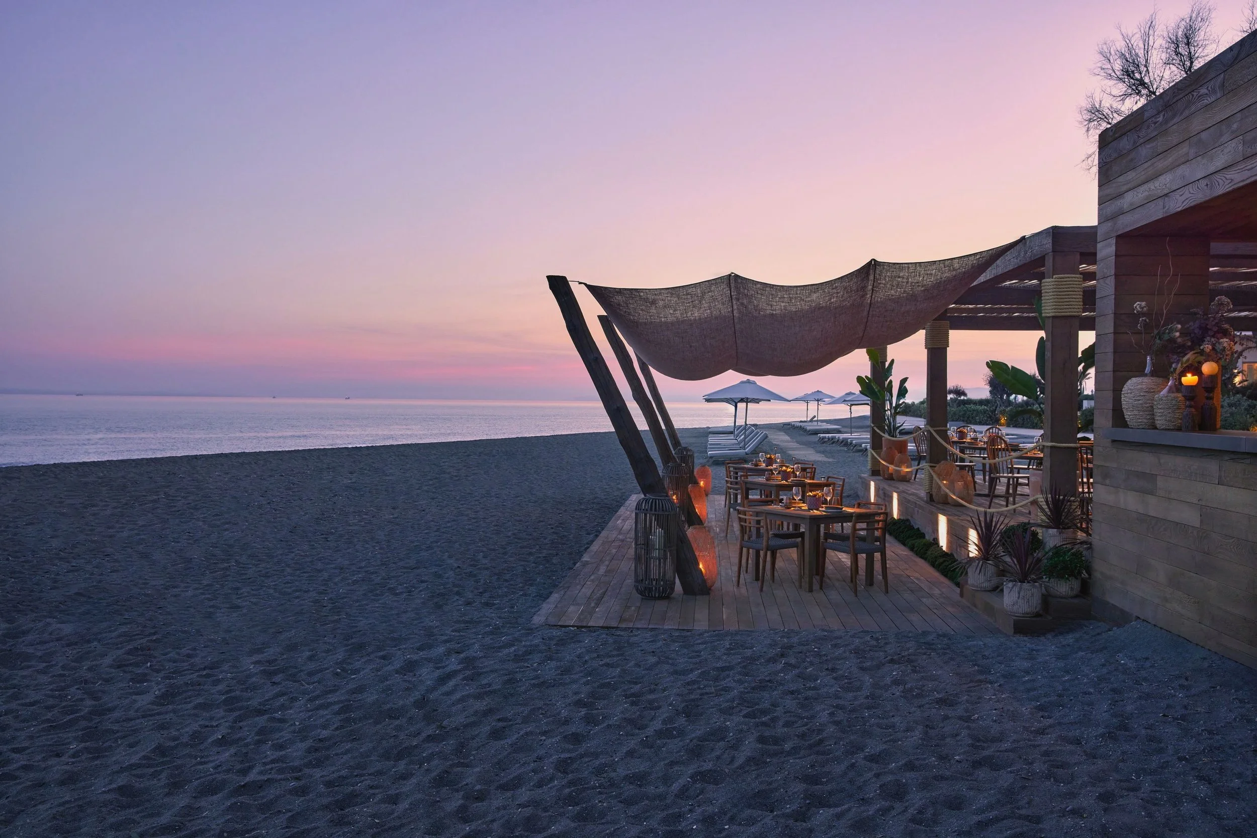 Beachside restaurant with outdoor seating area, string shade, and sunset view of the ocean.