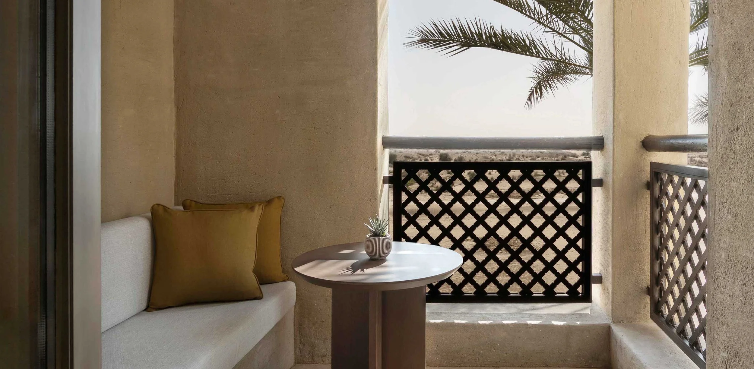 Small balcony with a white couch, yellow pillows, a round table, a potted plant, and a view of palm trees and desert landscape in the background.