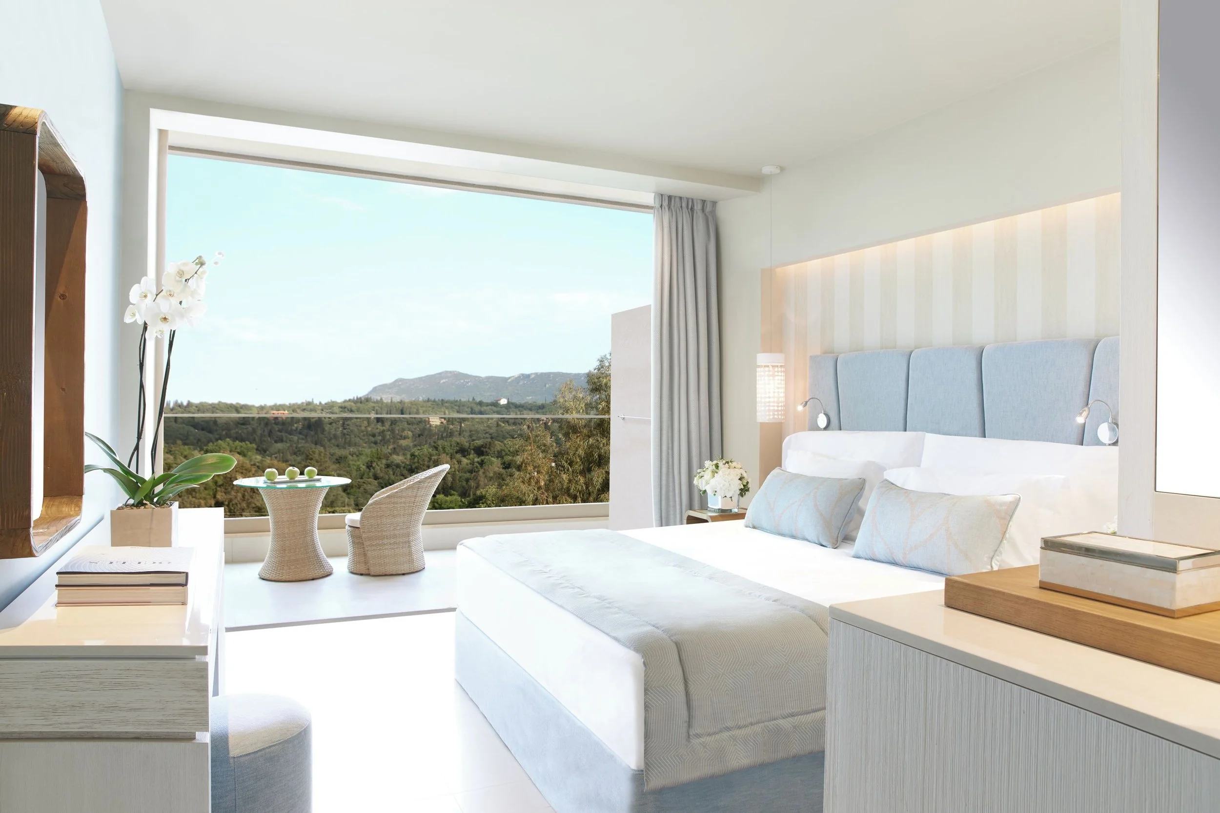 Bright hotel room with large window, view of green hills, and modern decor, including a bed with light blue headboard, white bedding, and small bedside lights.
