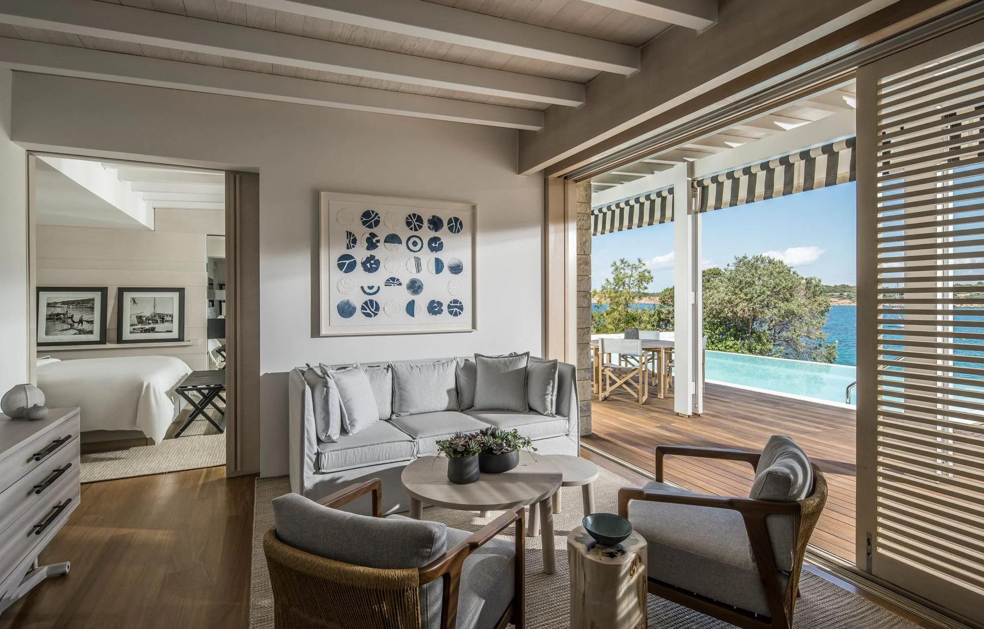 Living room with white sofa, armchair, and coffee table overlooking a patio with a pool and water view through open sliding glass doors, with striped awning outside.