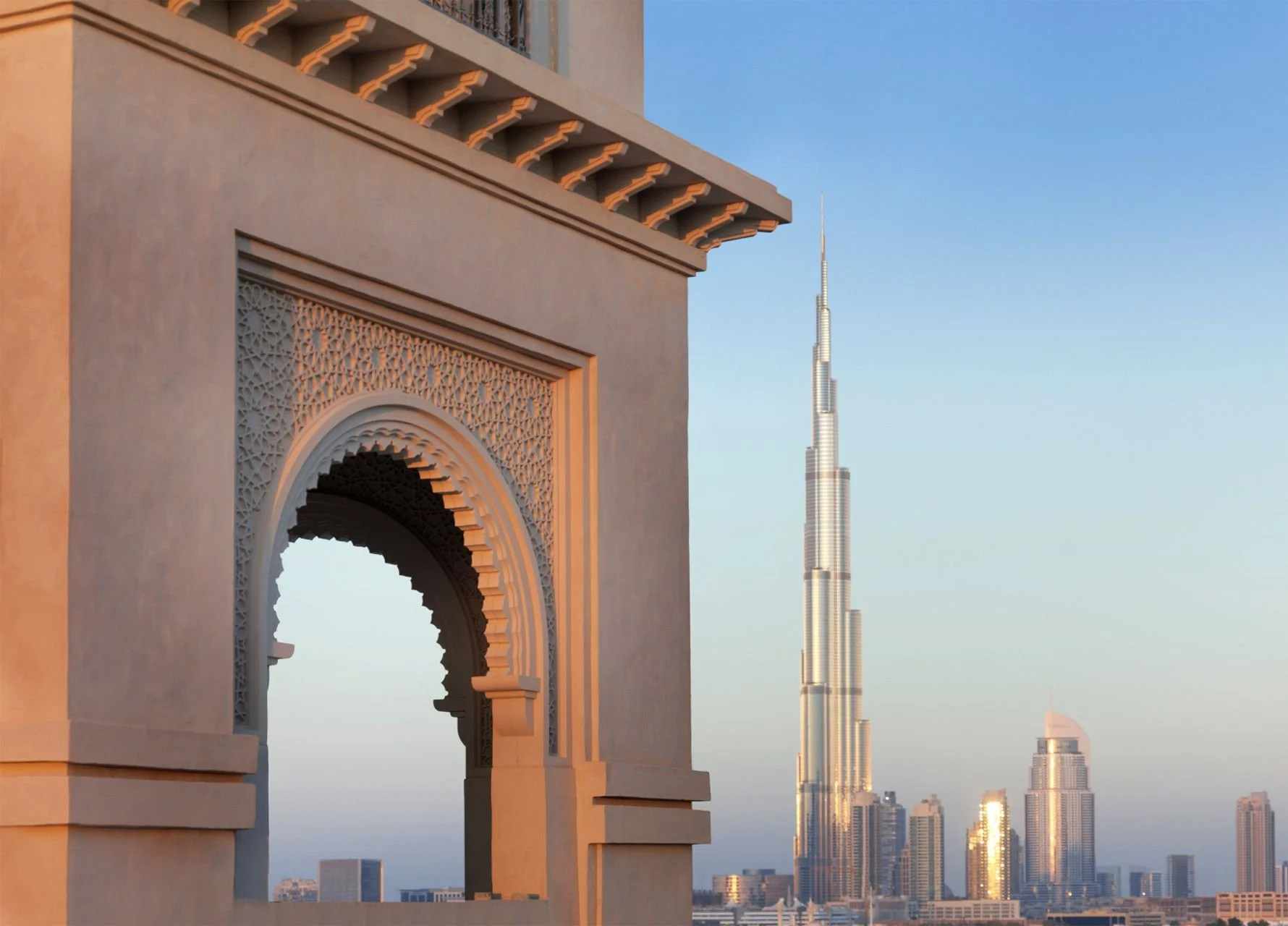 The image shows an ornate archway with detailed carvings on the left, overlooking a city skyline with the Burj Khalifa in Dubai in the background during sunset.