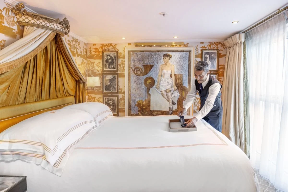 A hotel worker making a bed in a luxurious room with ornate gold and cream decor, gold curtains, and an abstract painting of a woman on the wall.