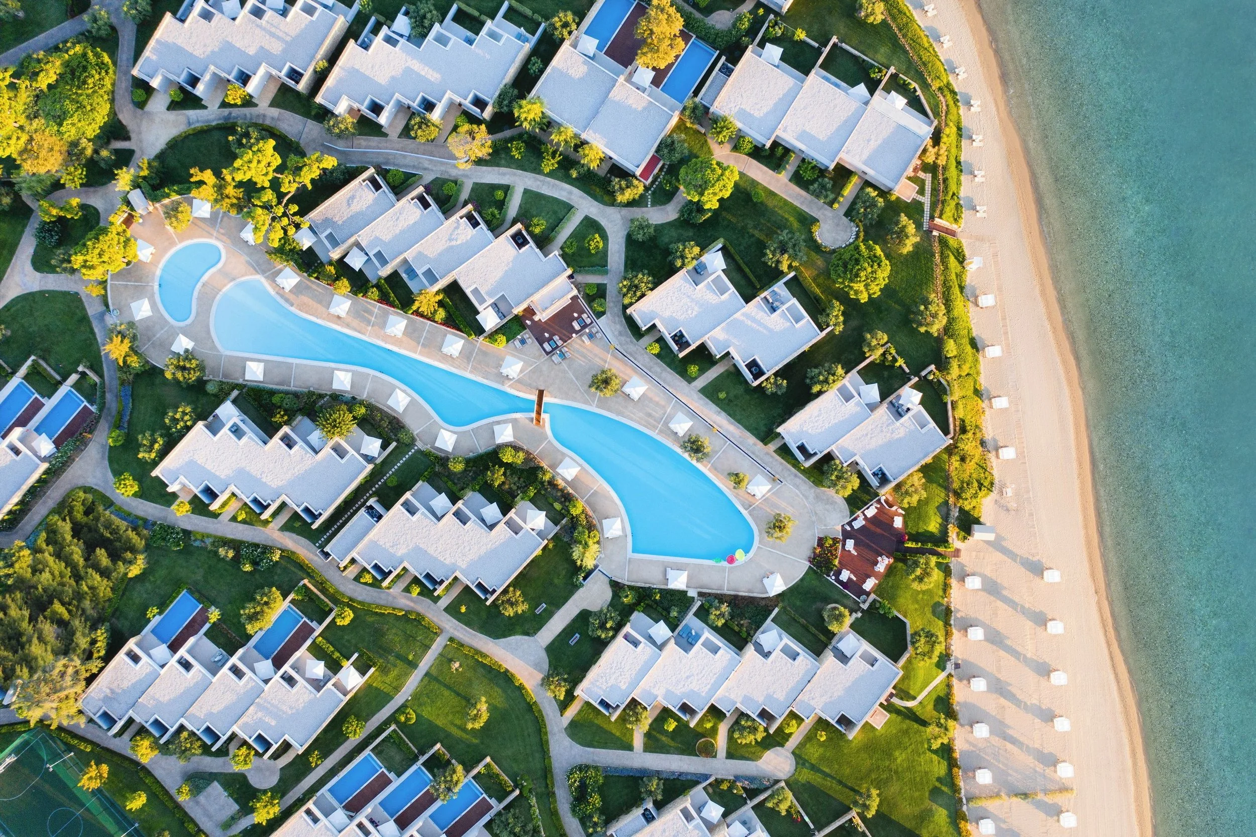  aerial view of a resort with multiple white-roofed buildings, winding pathways, a large swimming pool, green trees, and a beach along the shoreline.