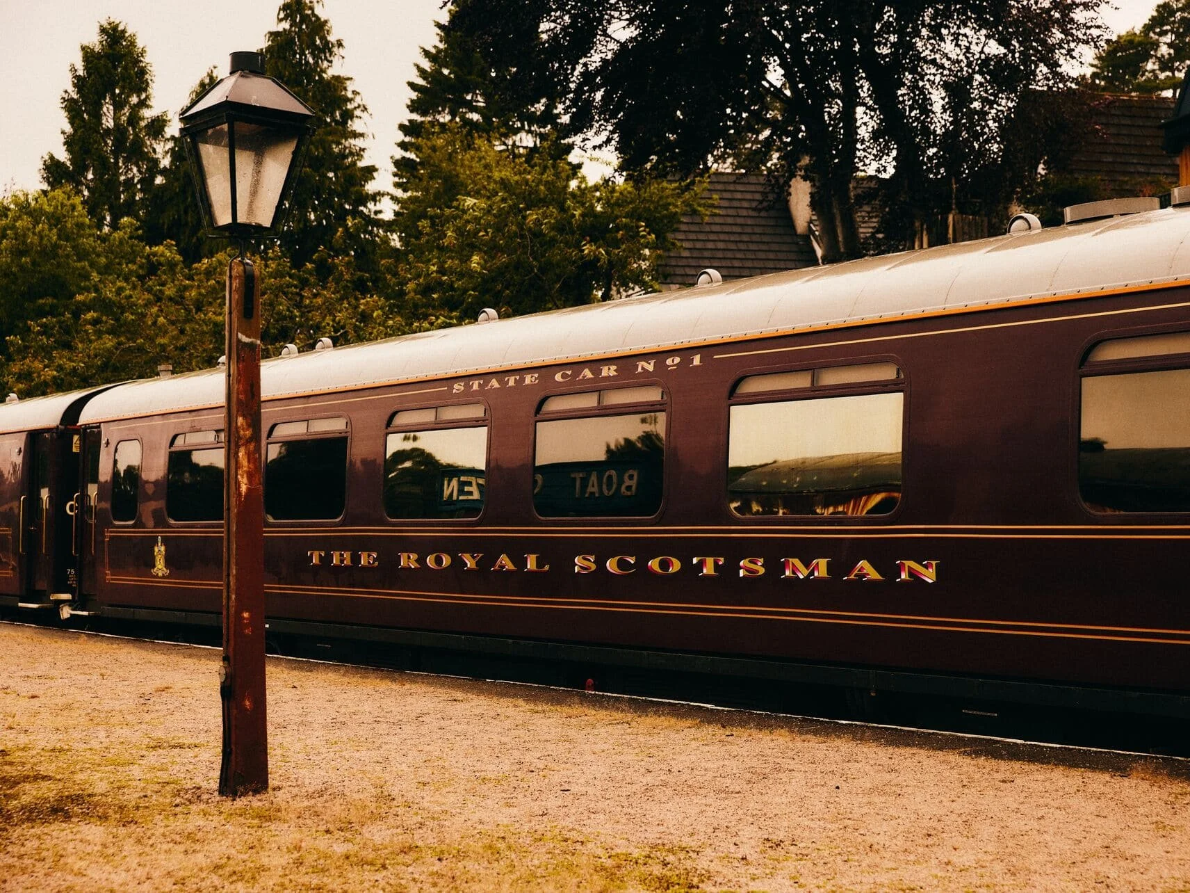 A vintage train car with the words 'The Royal Scotsman' in large gold and red letters and 'State Car No 1' in gold above the windows, parked outdoors with trees in the background and an old-fashioned street lamp in the foreground.
