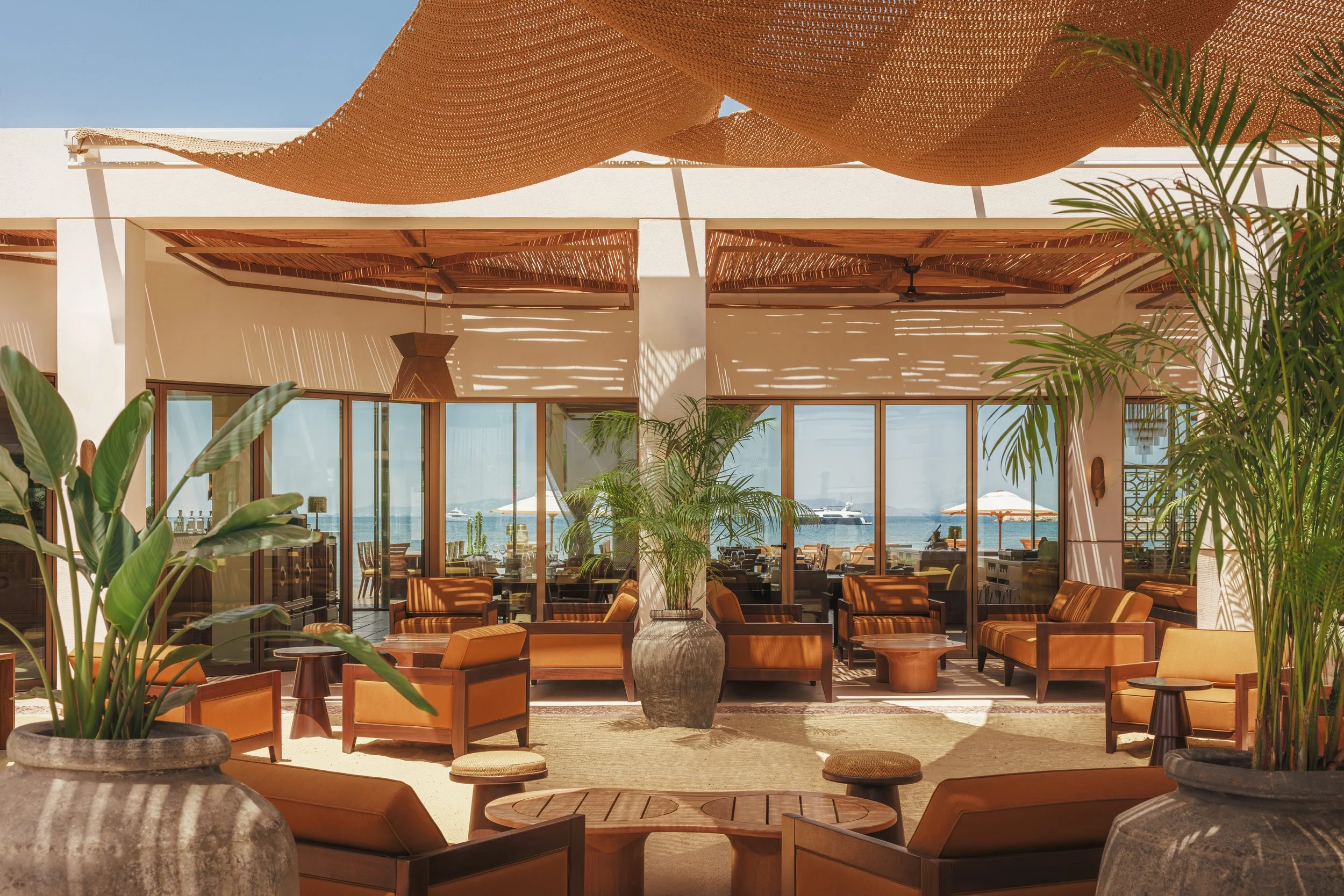 Interior of a beachside restaurant with wooden furniture, large potted plants, and a view of the ocean through glass windows.