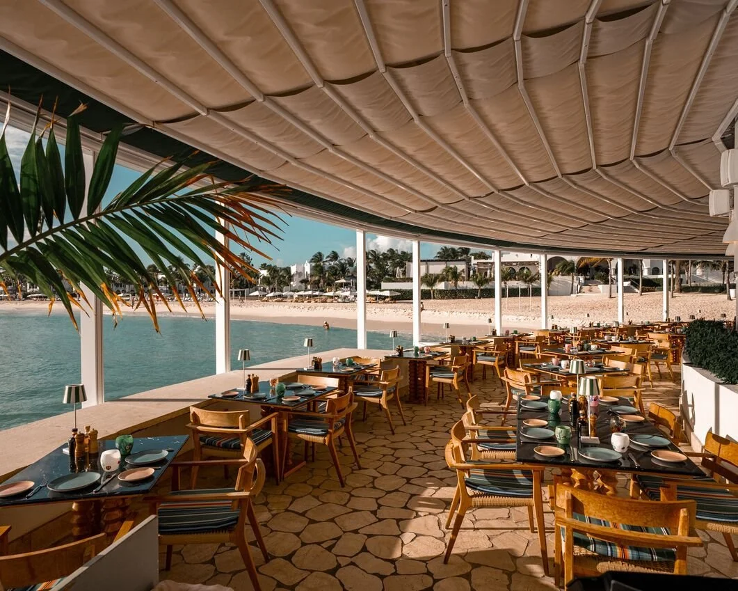 A beachside restaurant with tables set for dining, outdoor view of sandy beach and calm water, palm trees in the distance, and a partly shaded ceiling with a fabric cover.