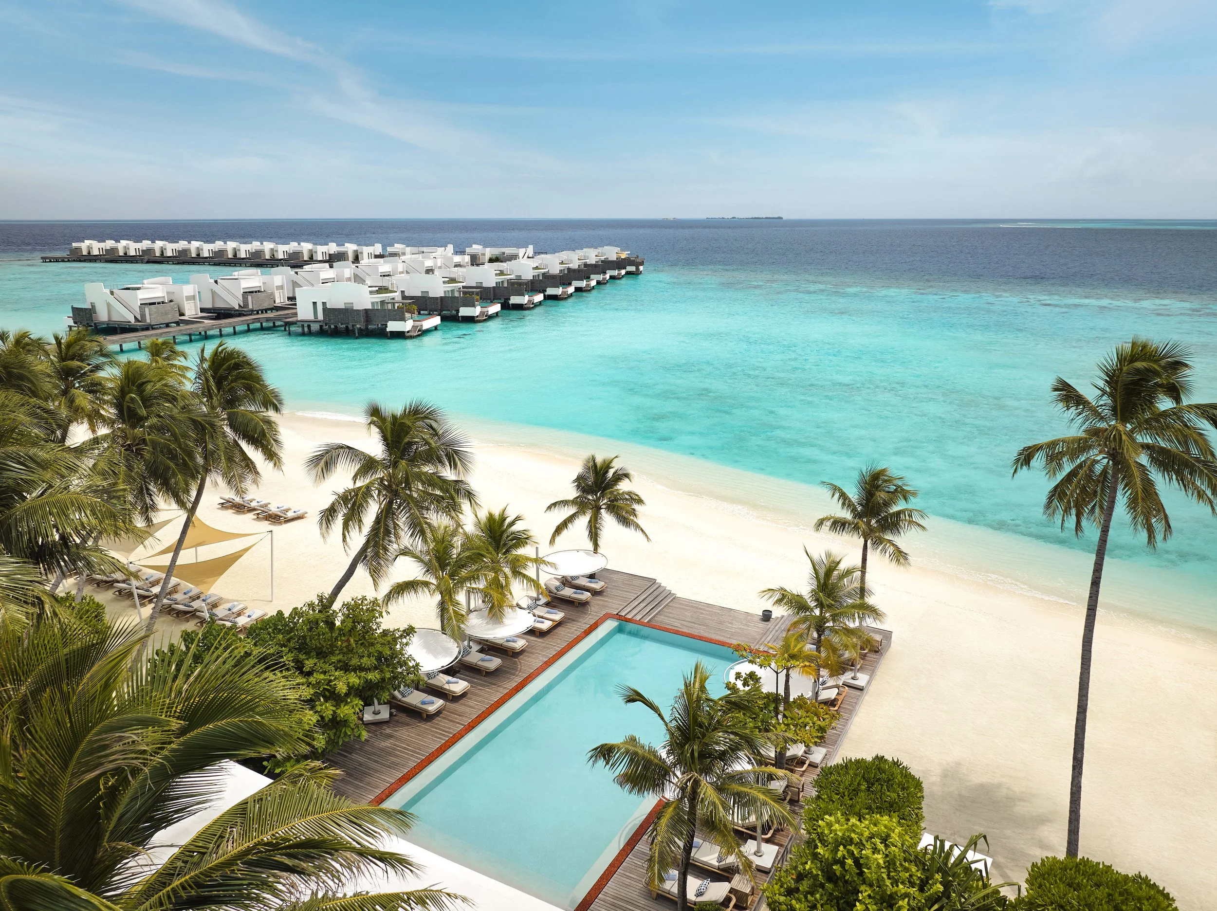 Aerial view of a luxury oceanfront resort with overwater bungalows, swimming pools, and palm trees on a sunny day with clear blue skies.