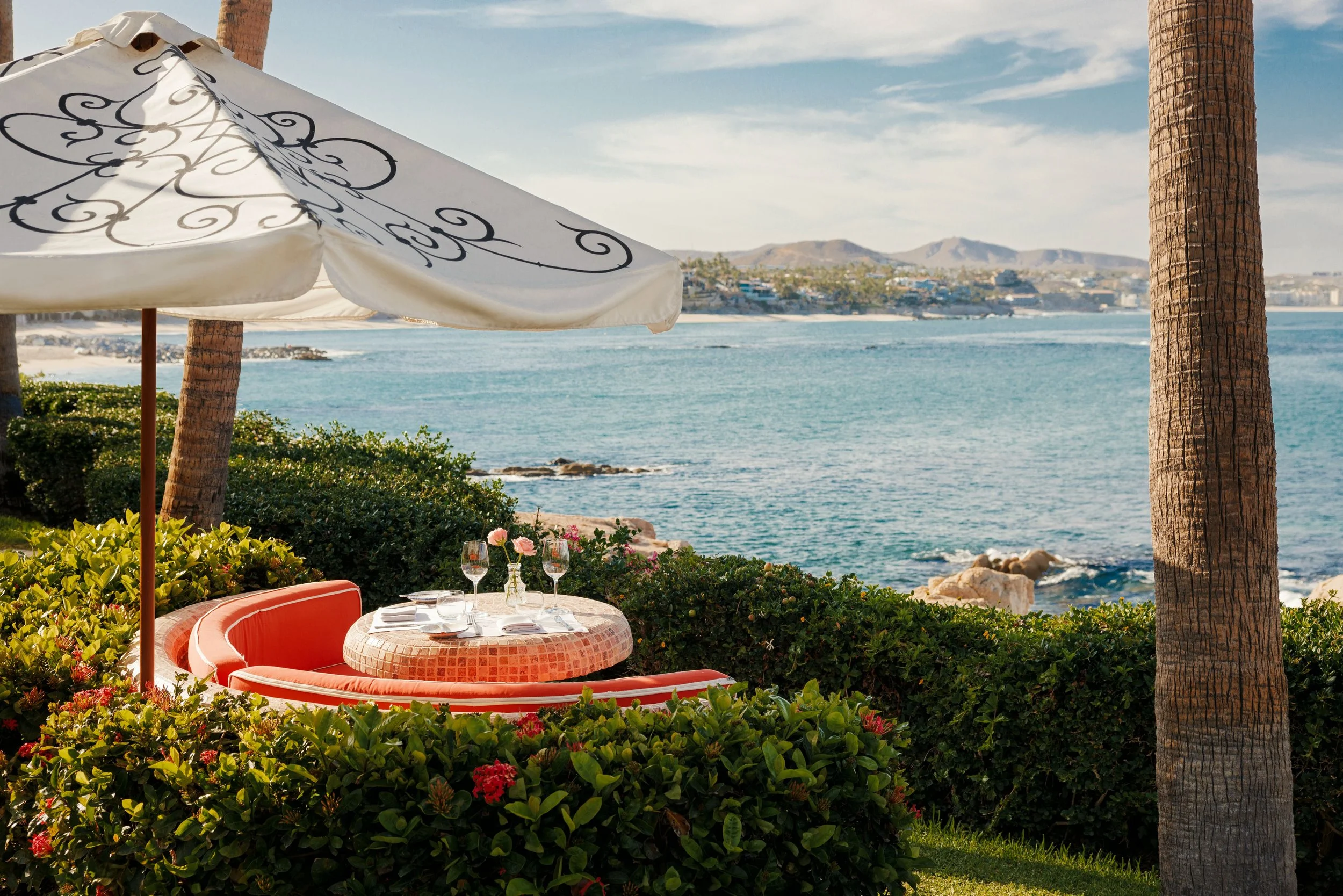 A seaside outdoor dining setup with a round table, orange cushioned bench, glasses, and plates, partially shaded by a large white umbrella with black decorative patterns, surrounded by lush greenery and flowering plants, overlooking the ocean with a distant view of mountains.