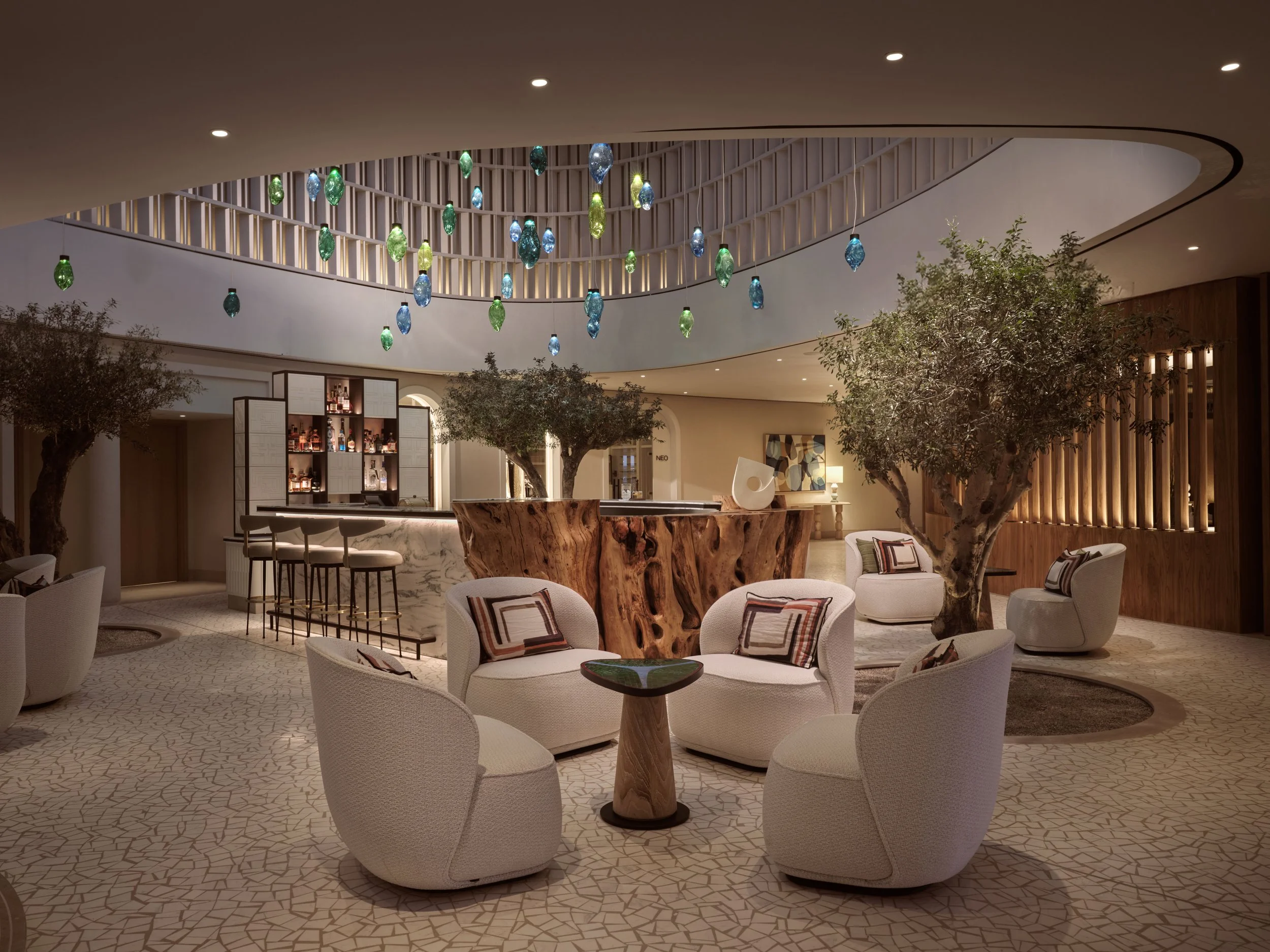 Interior of a modern lounge with white round chairs, a large wood table, tall plants, and colorful pendant lights hanging from the ceiling.