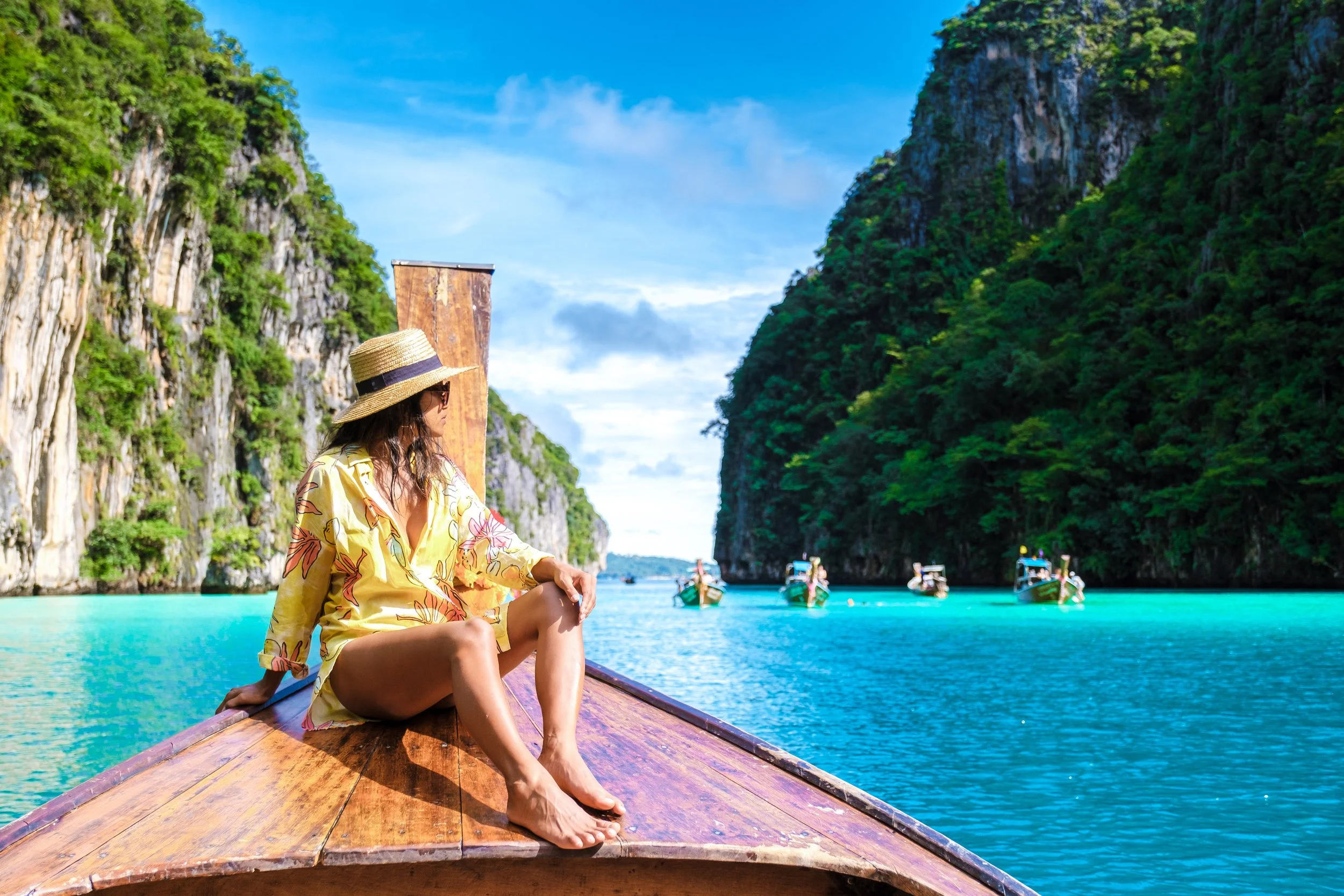 A woman sitting on the bow of a boat in a tropical bay with limestone cliffs covered in greenery, several boats in the water, and a bright blue sky.