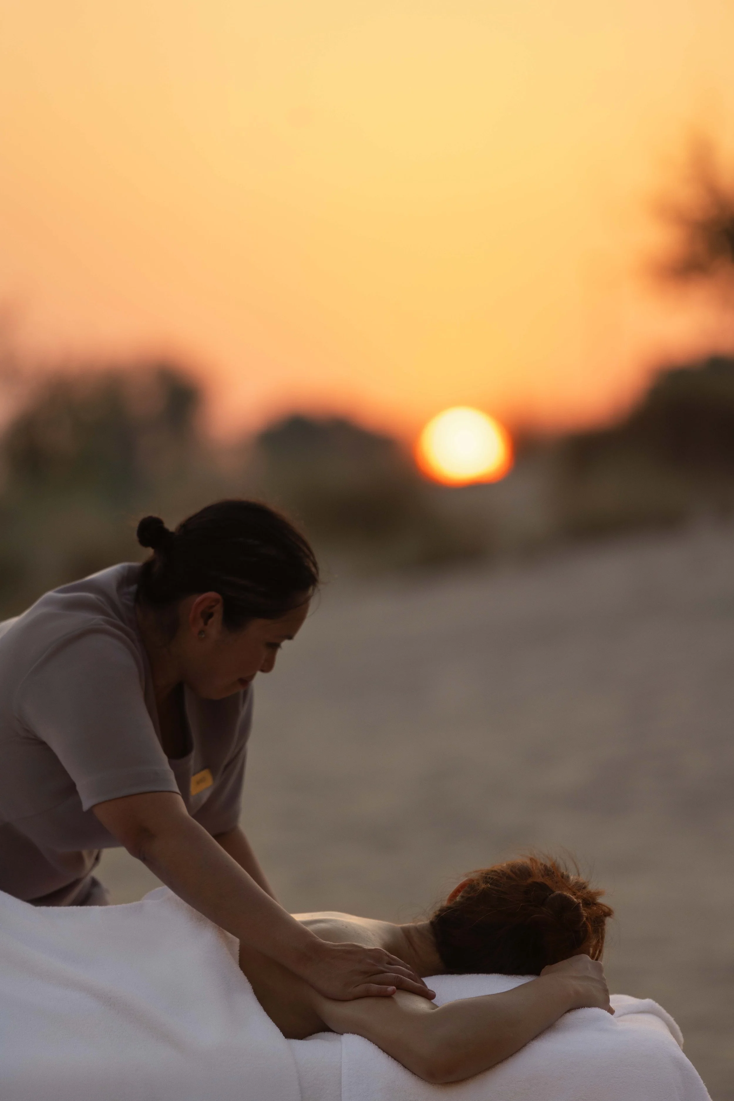 A woman receiving a massage on her back outdoors during sunset, with trees and a blurry sky in the background.
