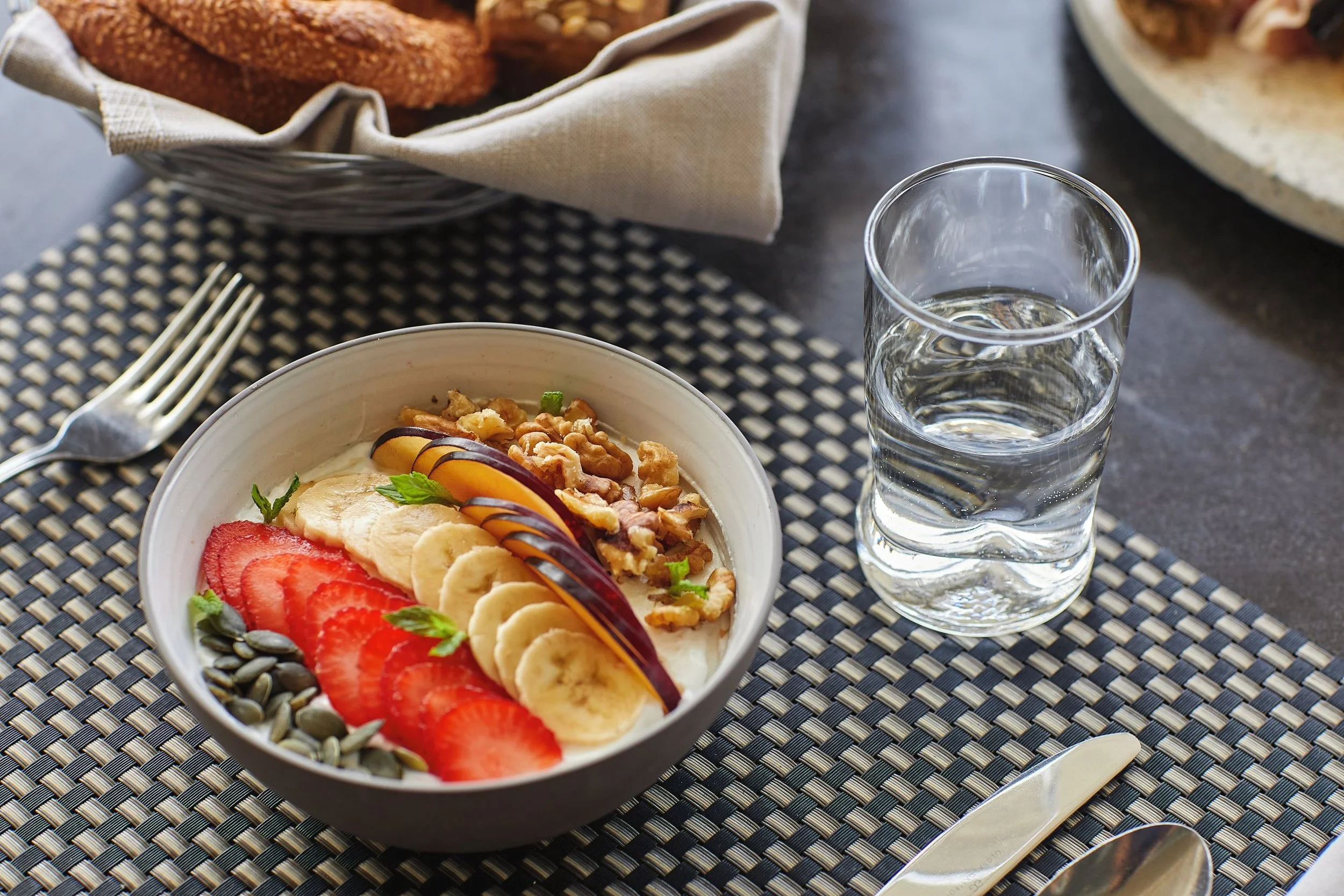 A bowl of yogurt topped with sliced strawberries, bananas, and a sprinkle of seeds, sitting on a black and white woven placemat; a glass of water and a fork are next to it, with a basket of bread in the background.