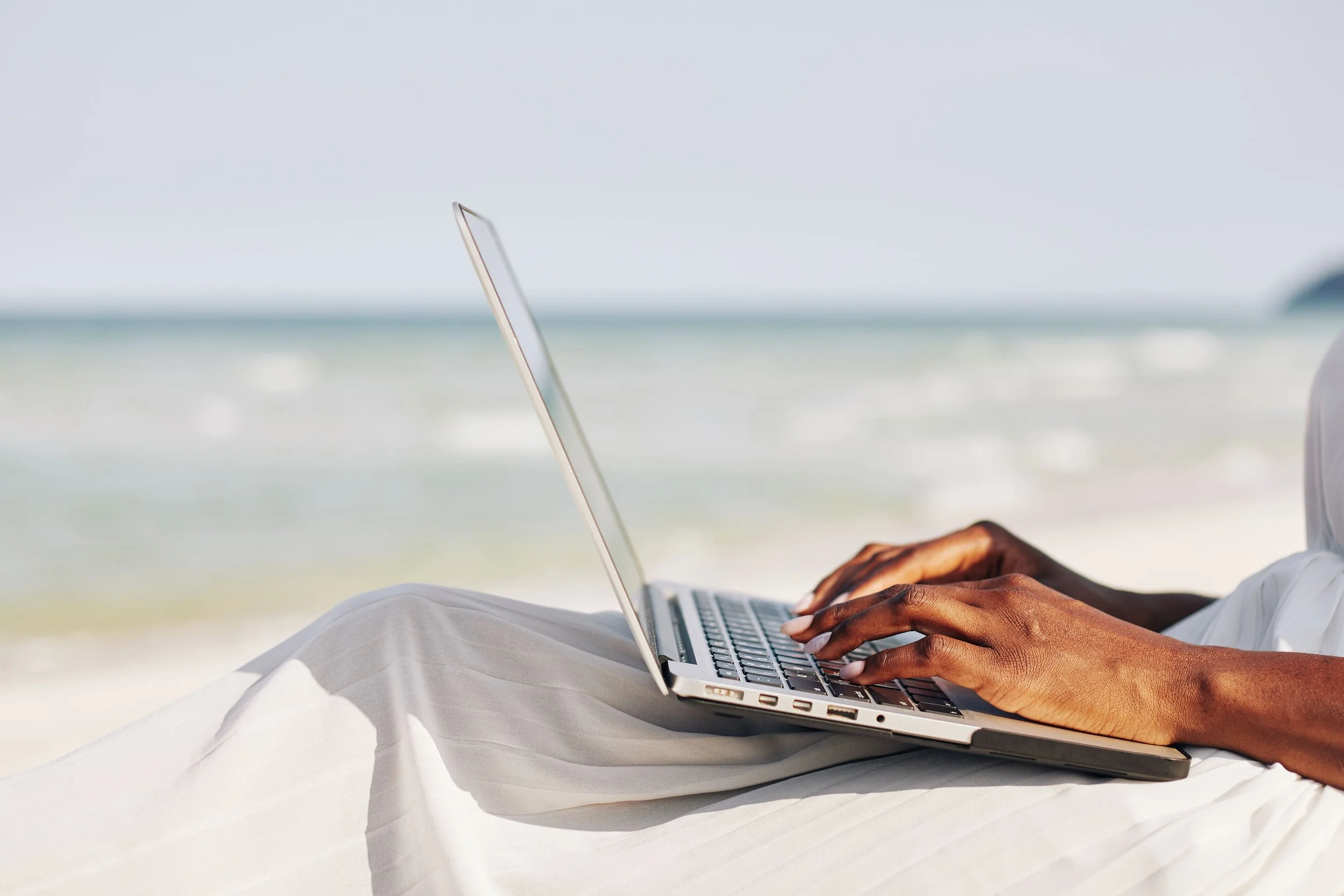 A person with dark skin sitting on a beach while using a laptop, with the ocean in the background.