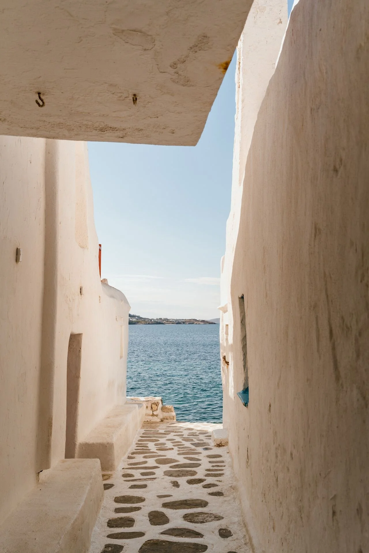 A narrow stone pathway between white plastered buildings leading to a view of a body of water, with a distant landmass and blue sky in the background.
