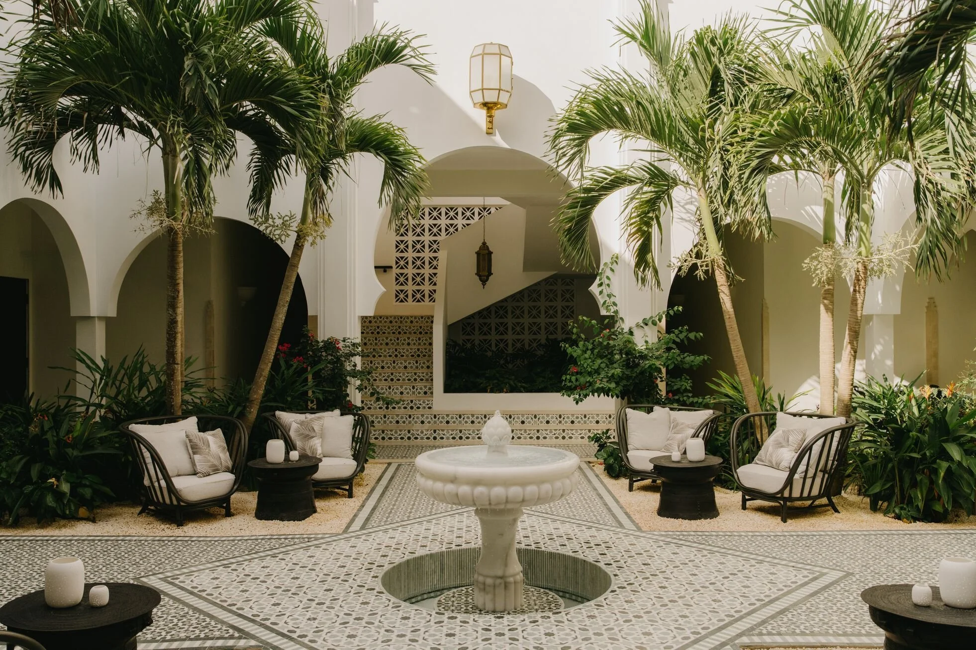 Indoor courtyard with white walls, tall palm trees, and a decorative fountain in the center, surrounded by chairs with cushions and small side tables.