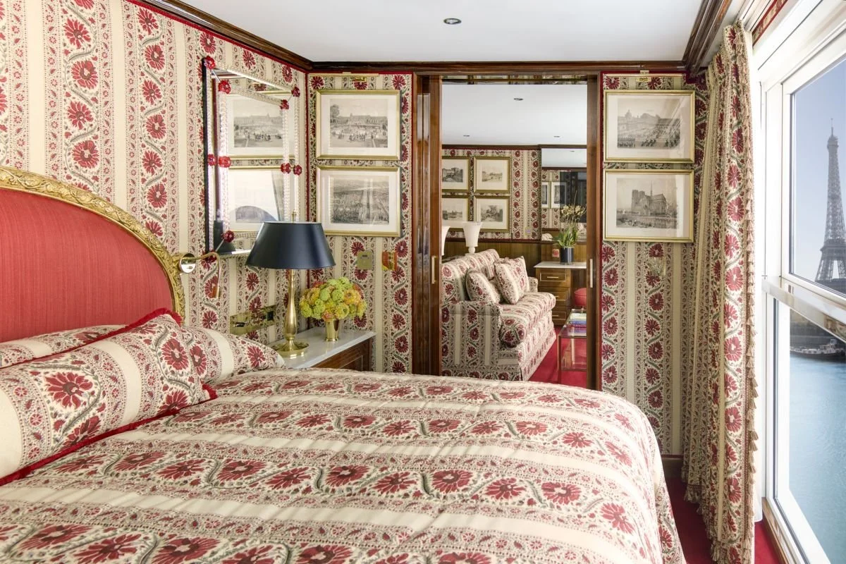 A bedroom with floral wallpaper, a large bed with a red and cream floral patterned bedspread, a matching headboard, a nightstand with a black lamp and a yellow flower arrangement, framed pictures on the wall, a mirror, and a window through which the Eiffel Tower is visible.