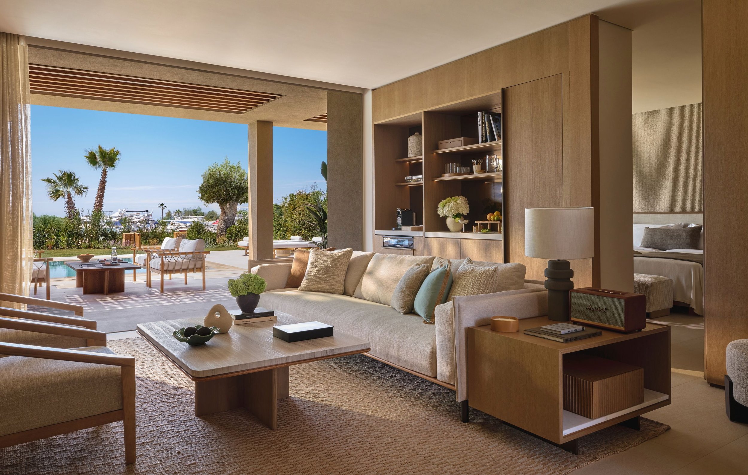 Living room with beige couch, wooden coffee table, and view of outdoor patio with white chairs, greenery, palm trees, and boats in distance.