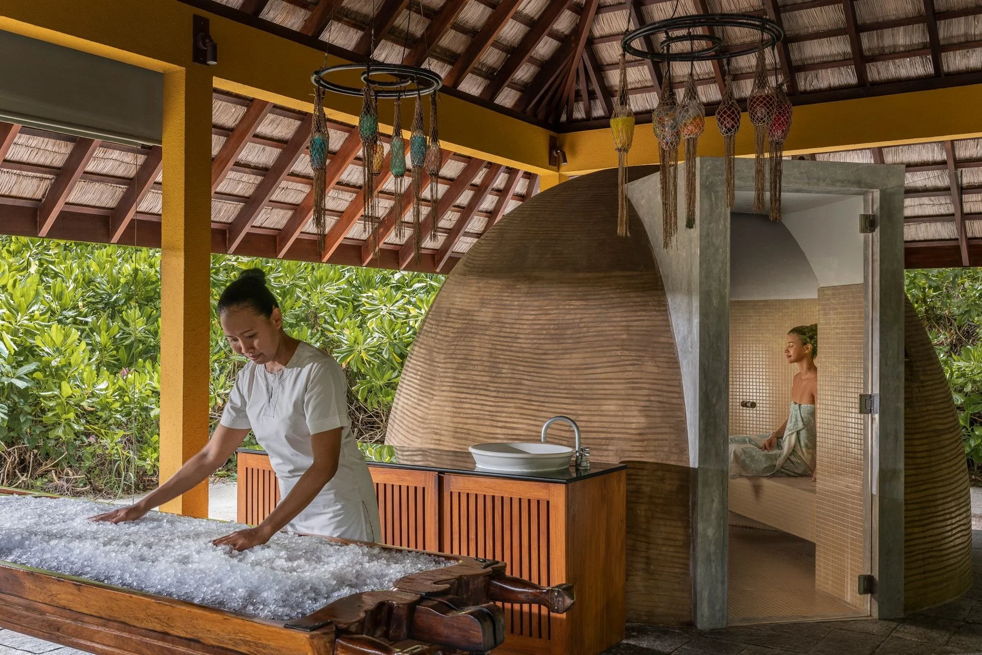 Woman in white shirt washing a textured surface, with a thatched roof, yellow wall, decorative hanging lanterns, and lush green plants outside, and another woman relaxing in a tiled room.