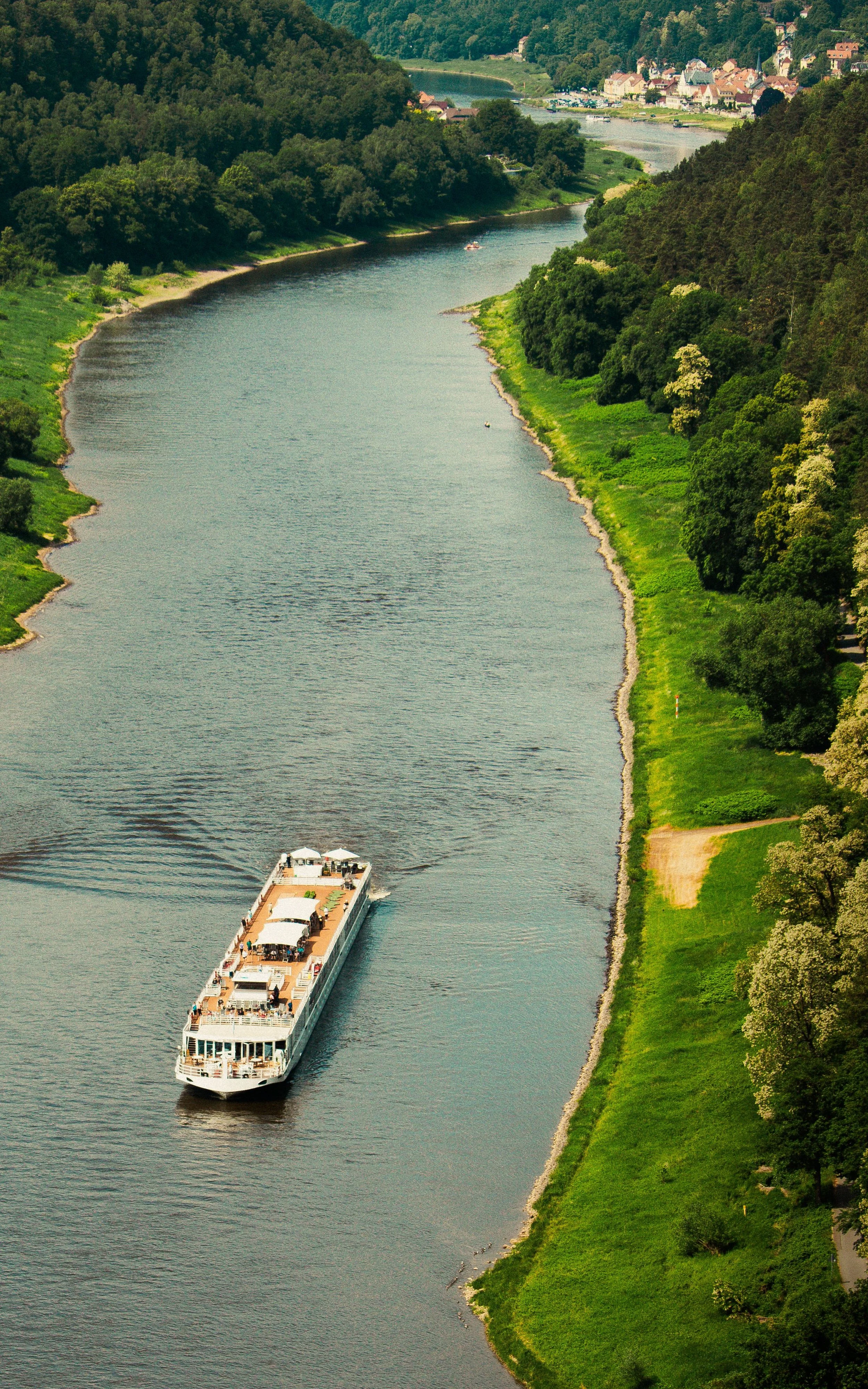 A river with a passenger cruise boat sailing along a lush green shoreline, under a sunny sky with a small town visible in the background.