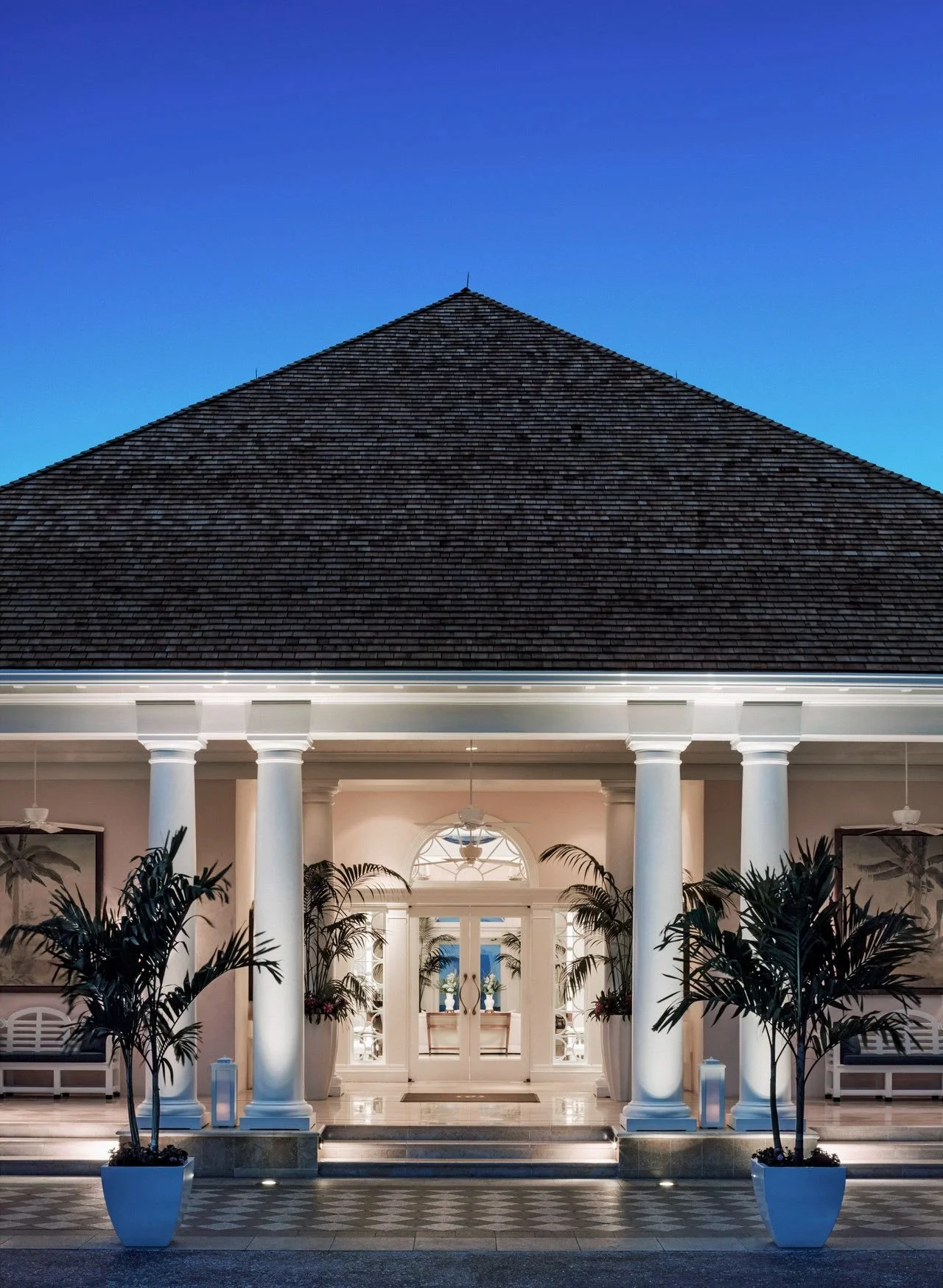 The image shows the front entrance of a house with a large, triangular shingled roof, white columns, indoor plants, and a glass-paneled door, illuminated warmly against a dusky sky.