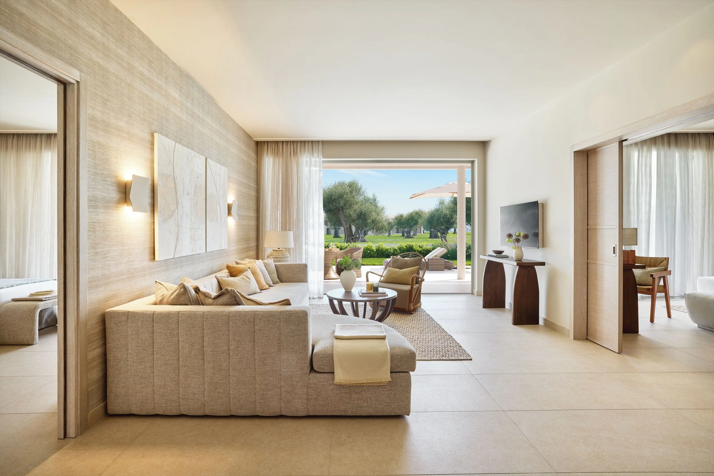 A spacious living room with a beige sofa, cream-colored armchair, and coffee table with a vase, overlooking a patio and a lush green landscape outside through large glass doors.