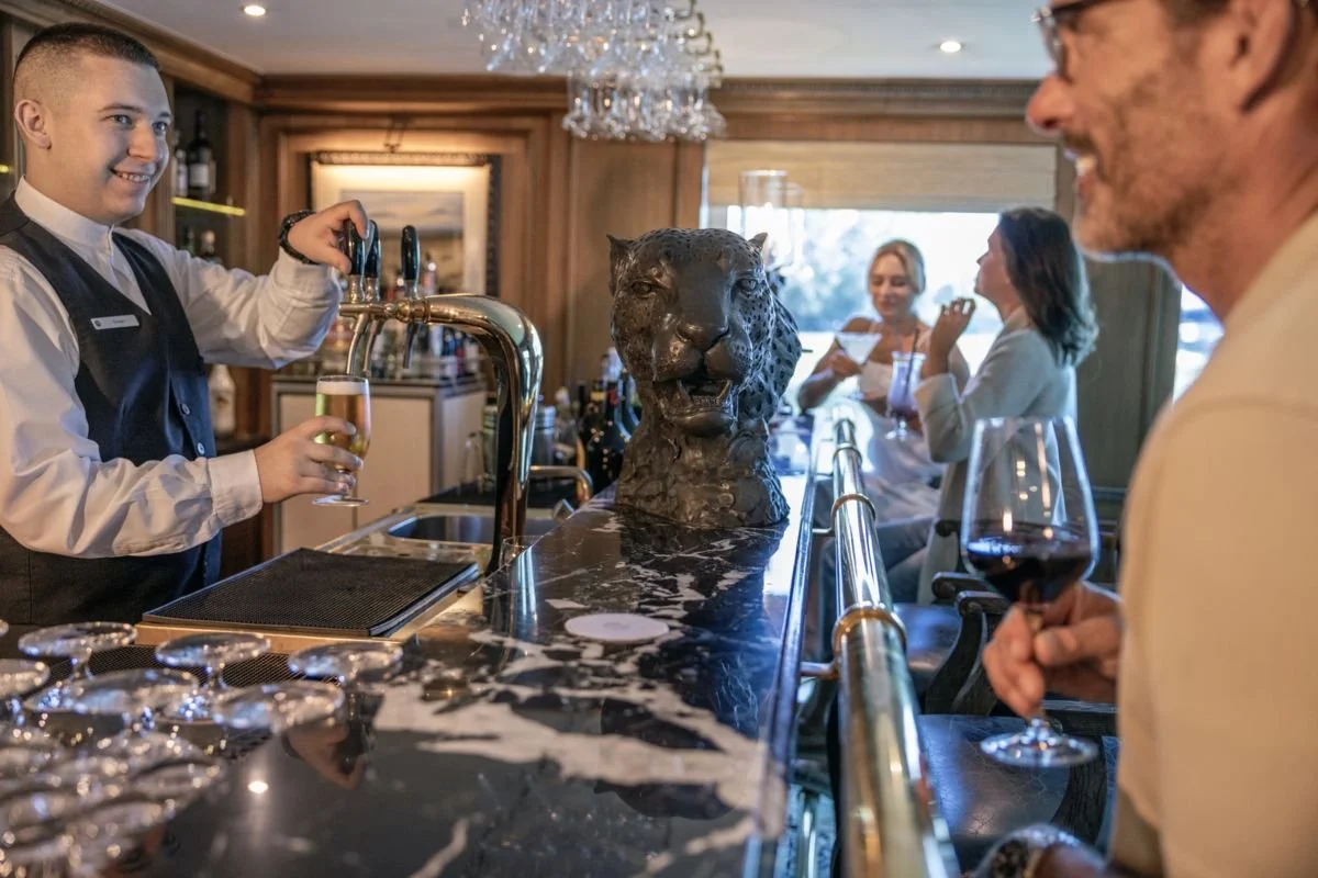 A bartender pouring a drink at a bar with adults enjoying wine and a decorative leopard sculpture on the counter.