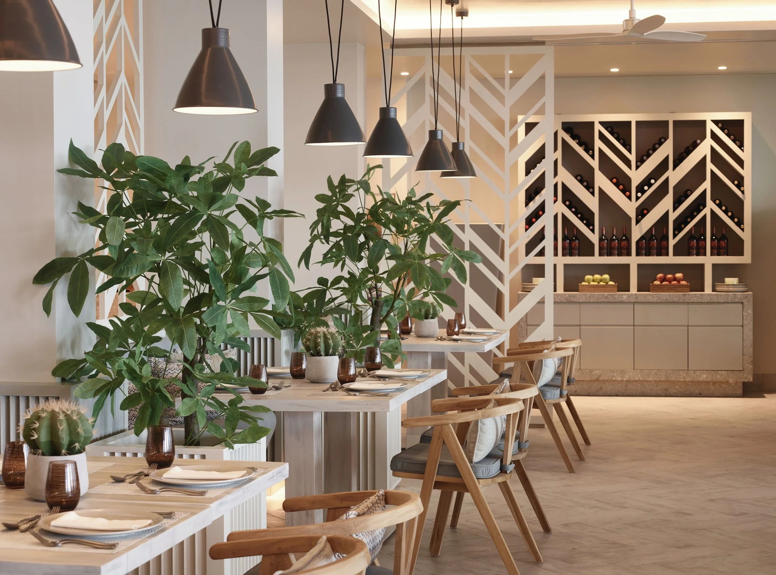 A modern restaurant interior with wooden tables set for dining, decorated with potted cacti and leafy green plants. Overhead, black pendant lights hang in a line. There's a white decorative room divider with a geometric pattern, and a black wine rack on the wall in the background.
