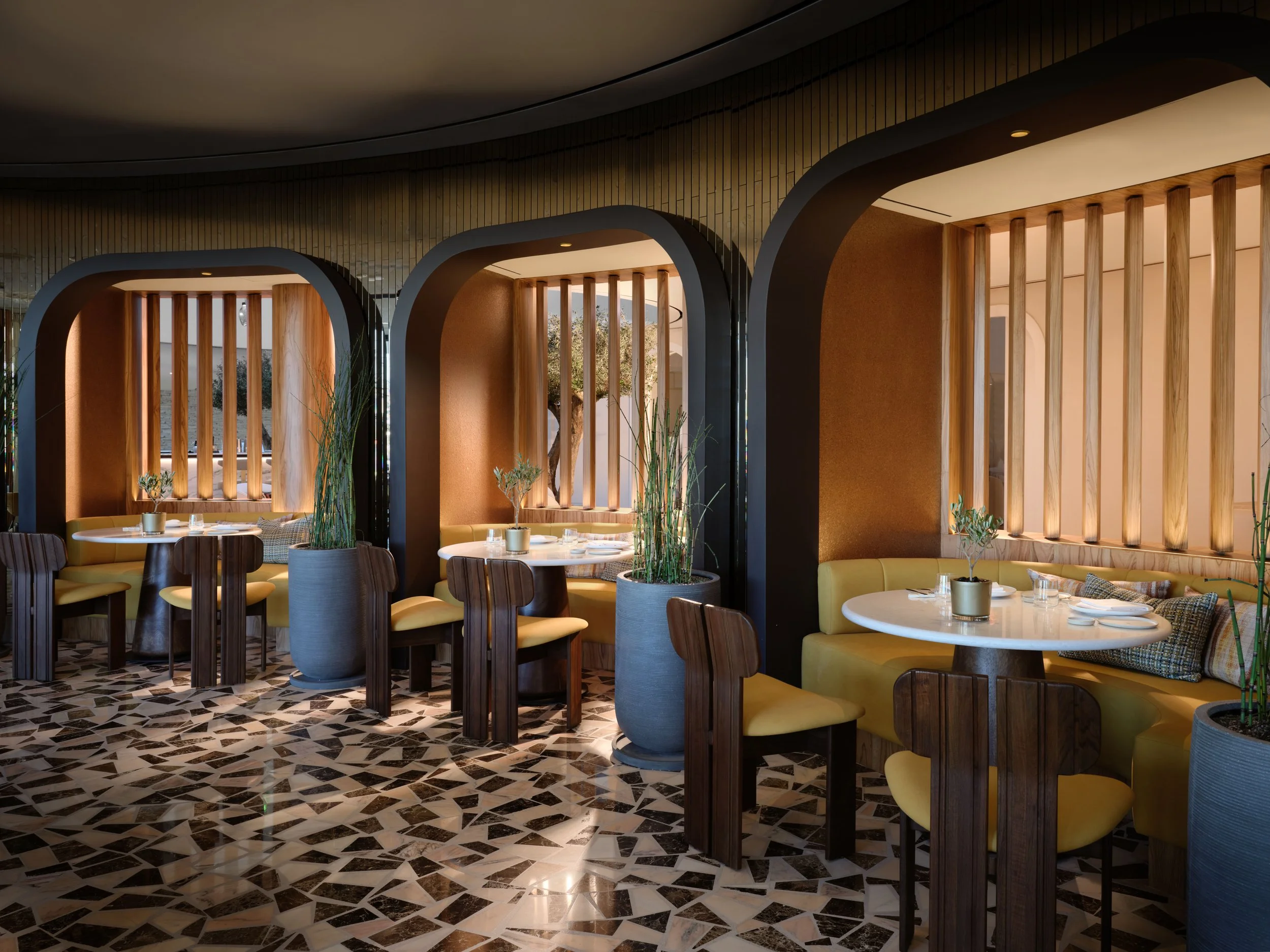 Interior of a modern restaurant with circular tables, yellow cushioned seating, wooden dividers, and a geometric black and white tiled floor.