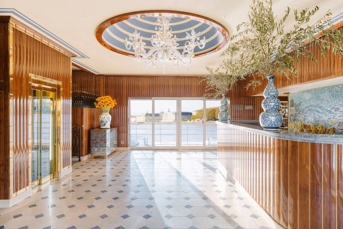 Elegant interior space with wooden paneling, a chandelier, large windows overlooking water, and decorative vases with flowers and branches.